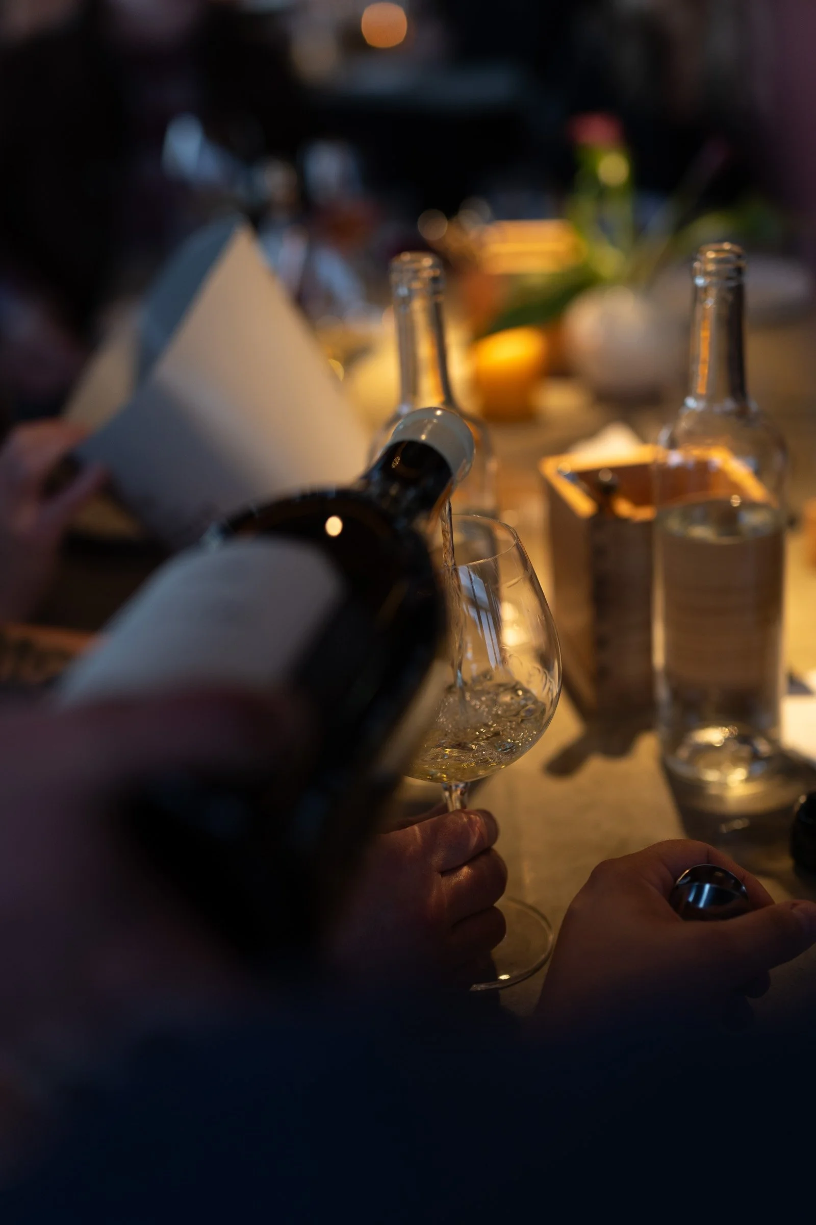 Person som heller hvitvin i et vinglass på et bord med flere tomme glass, vannflasker og lys i bakgrunnen.