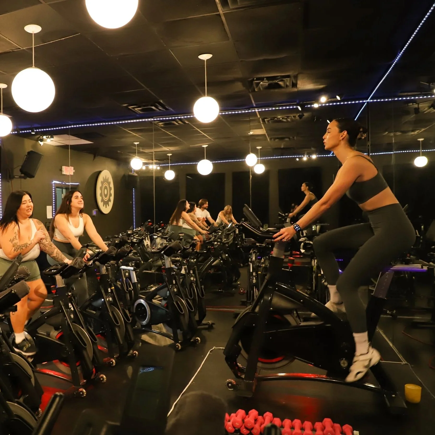 A group of women participating in a spin class in a dark, well-lit indoor gym, with one woman in the foreground on a stationary bike.