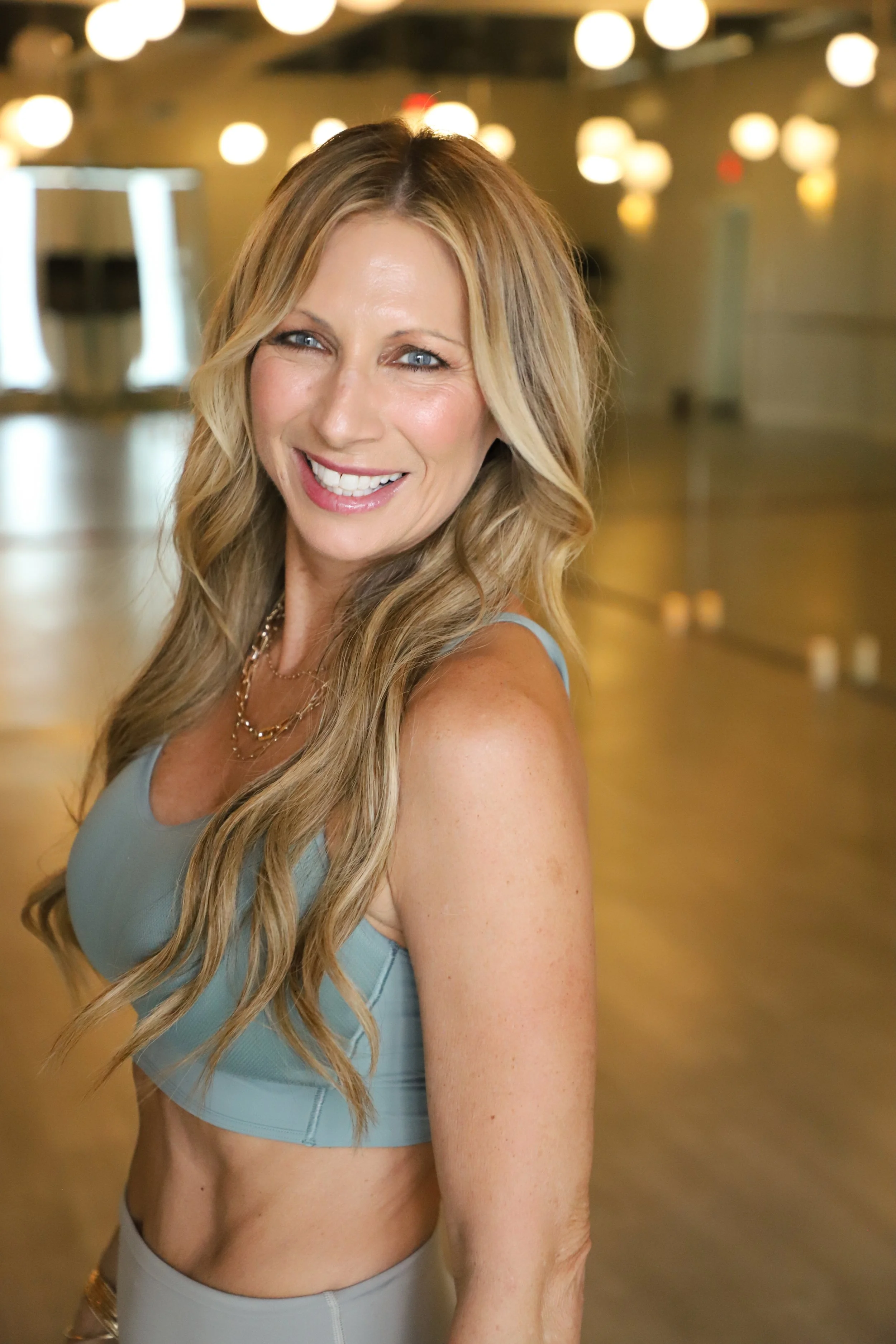 A smiling woman with long, wavy blonde hair in a fitness studio, wearing a light blue sports bra and beige workout pants.