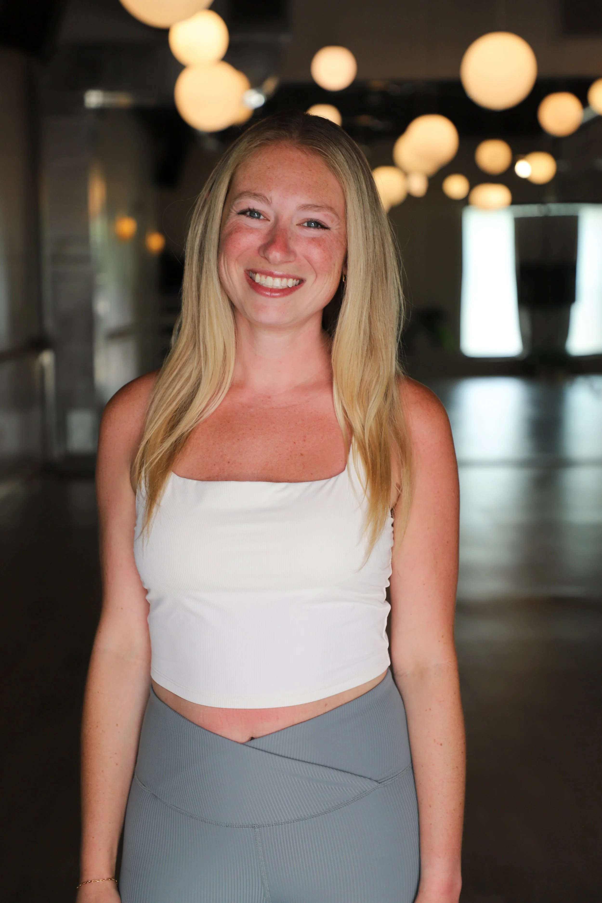 A smiling woman with long blonde hair, wearing a white sleeveless top and gray high-waisted leggings, standing indoors with warm round lights hanging from the ceiling in the background.