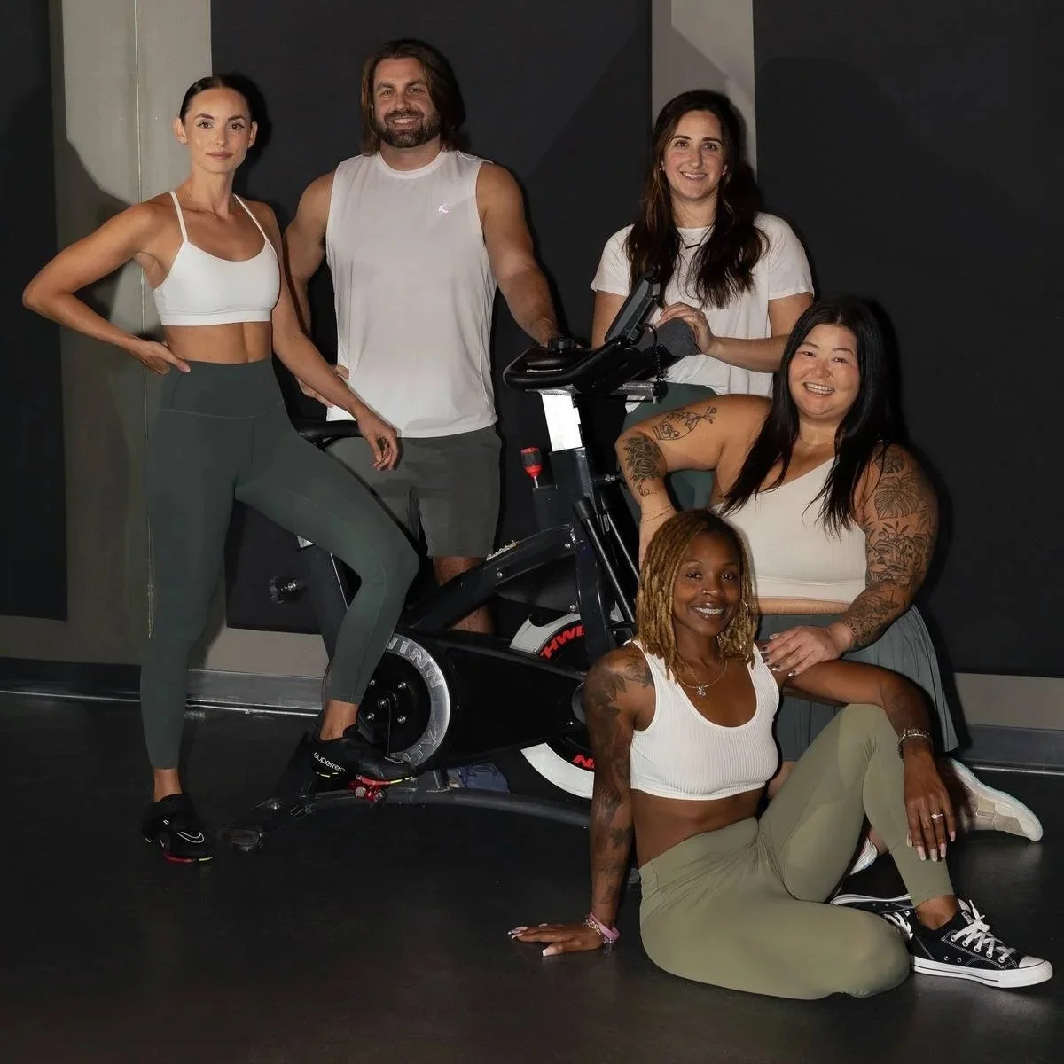 Group of six diverse women and one man in a fitness studio, with one woman using a stationary bike and others posing around her, smiling and showing fitness enthusiasm.