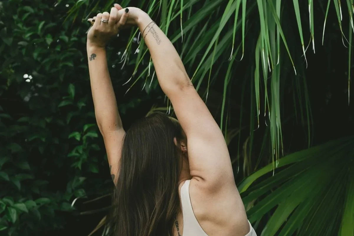 Woman with long brown hair stretching her arms over her head outdoors among green leafy plants.