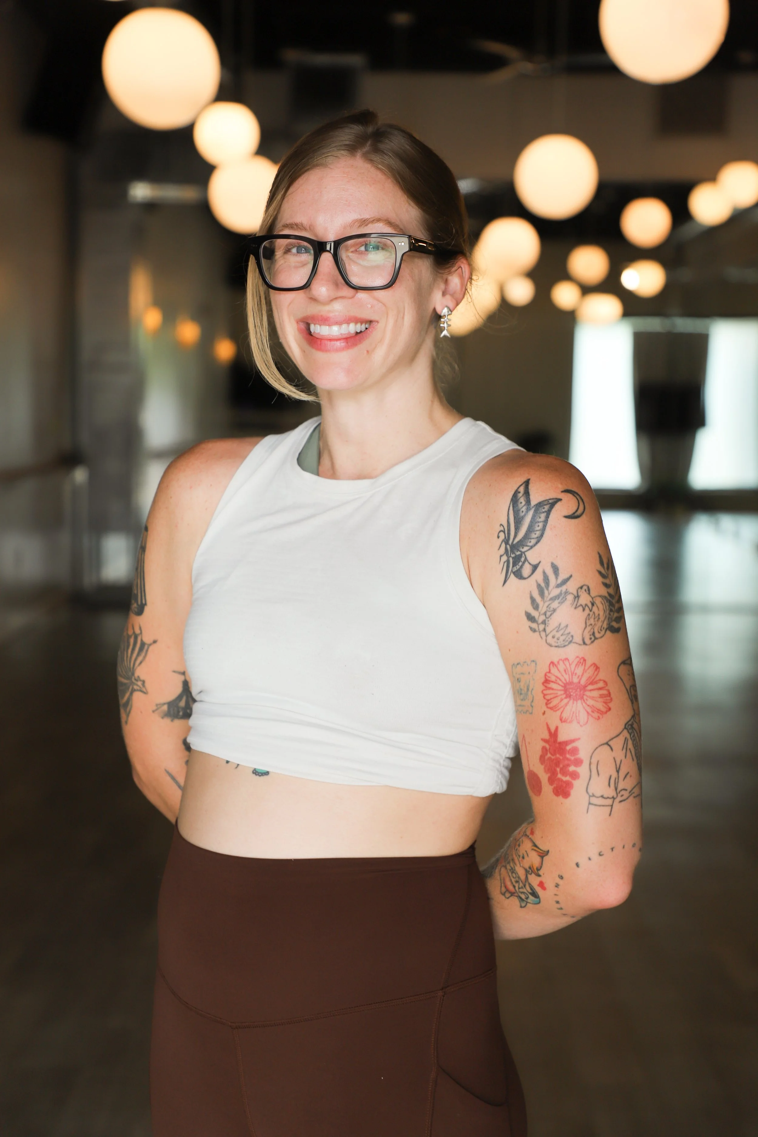 A woman wearing glasses and a sleeveless white crop top, smiling, with visible tattoos on her arms, standing in a room with round hanging lights.