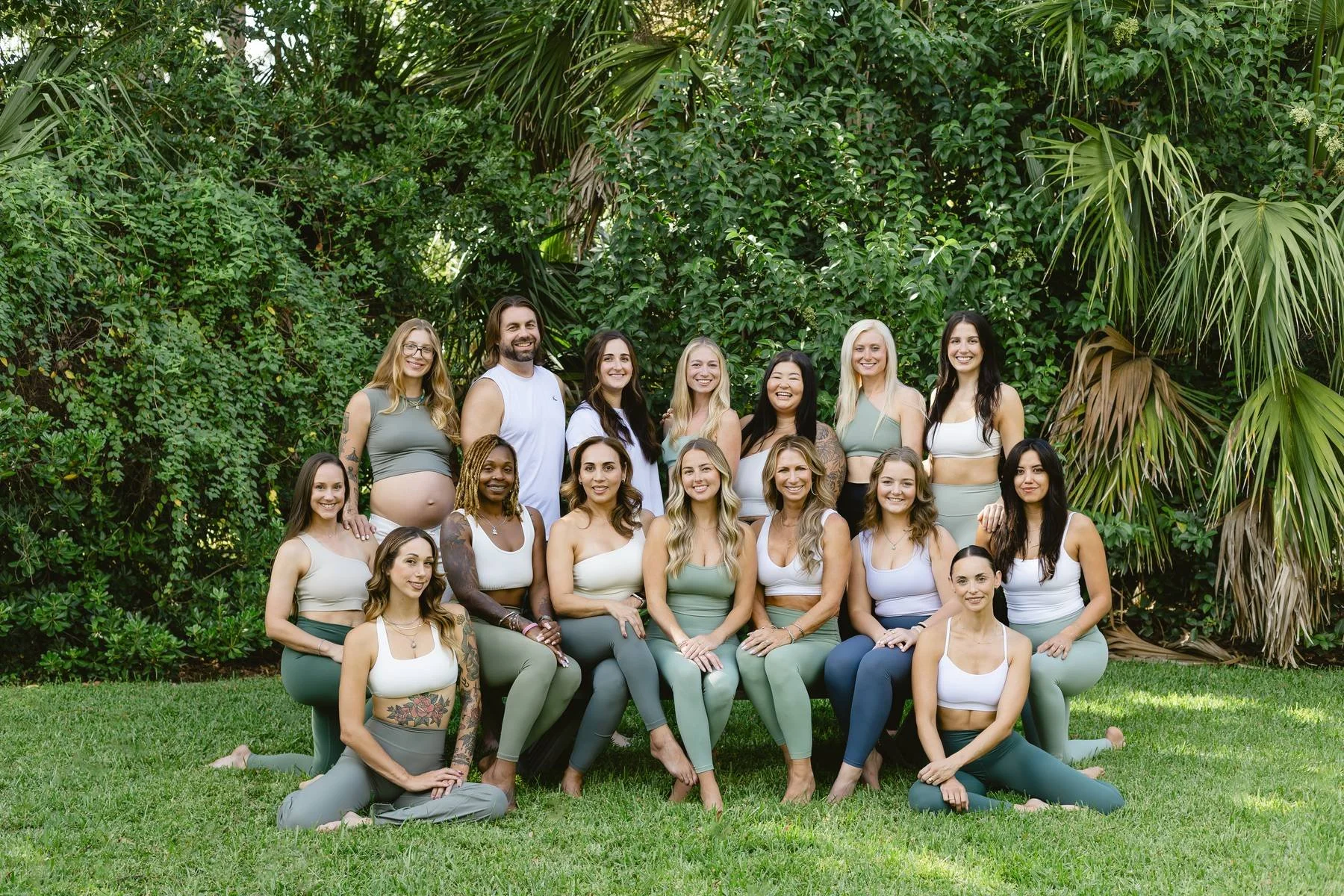 Group of women and one man posing outdoors on a grassy area with lush green plants and trees in the background.
