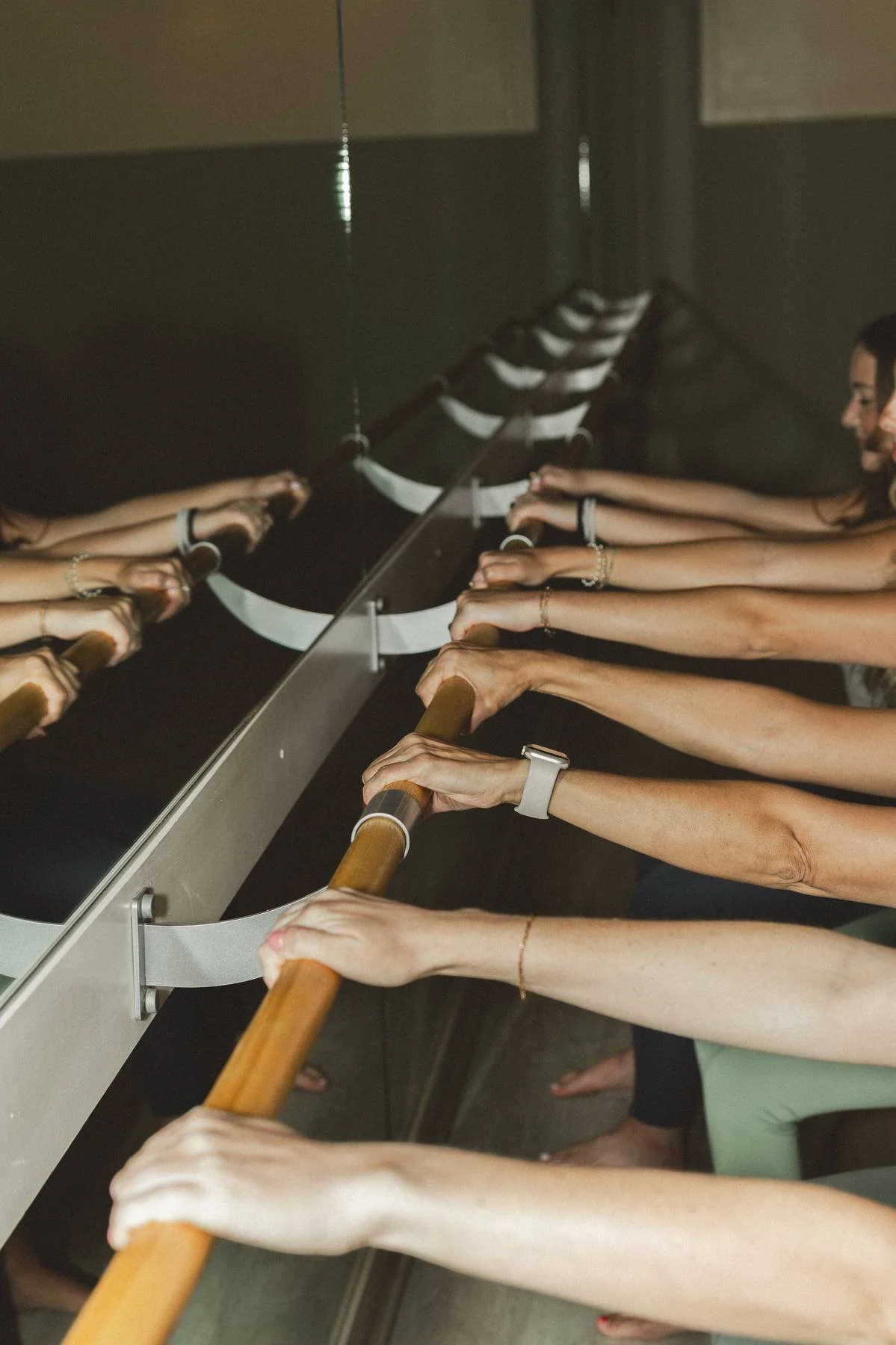 Several women doing a barre workout in front of a mirror, holding onto a wooden ballet barre with their hands extended forward.