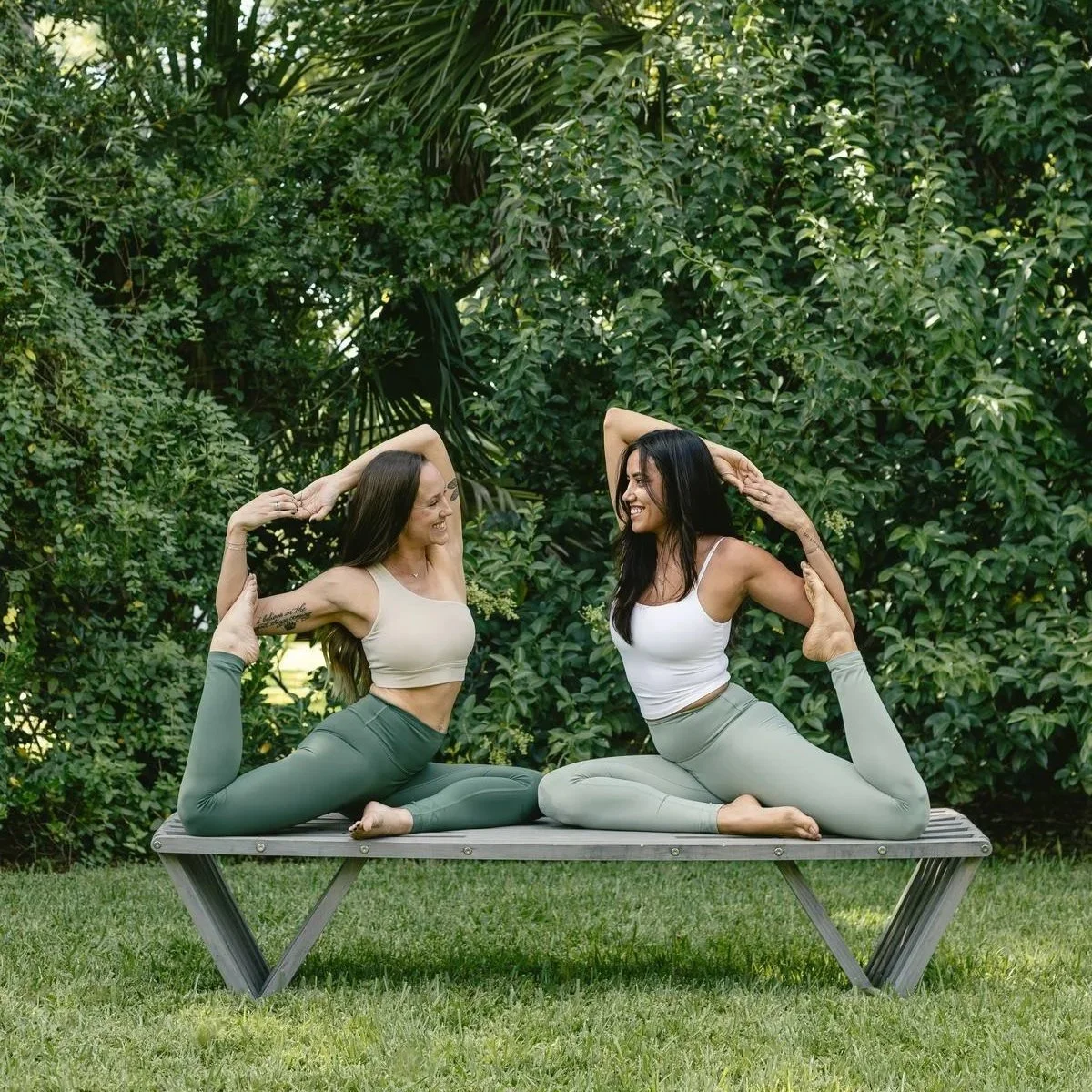 Two women practicing yoga outdoors on a wooden bench surrounded by lush green foliage, both smiling and in a backbend pose with arms raised and hands clasped.