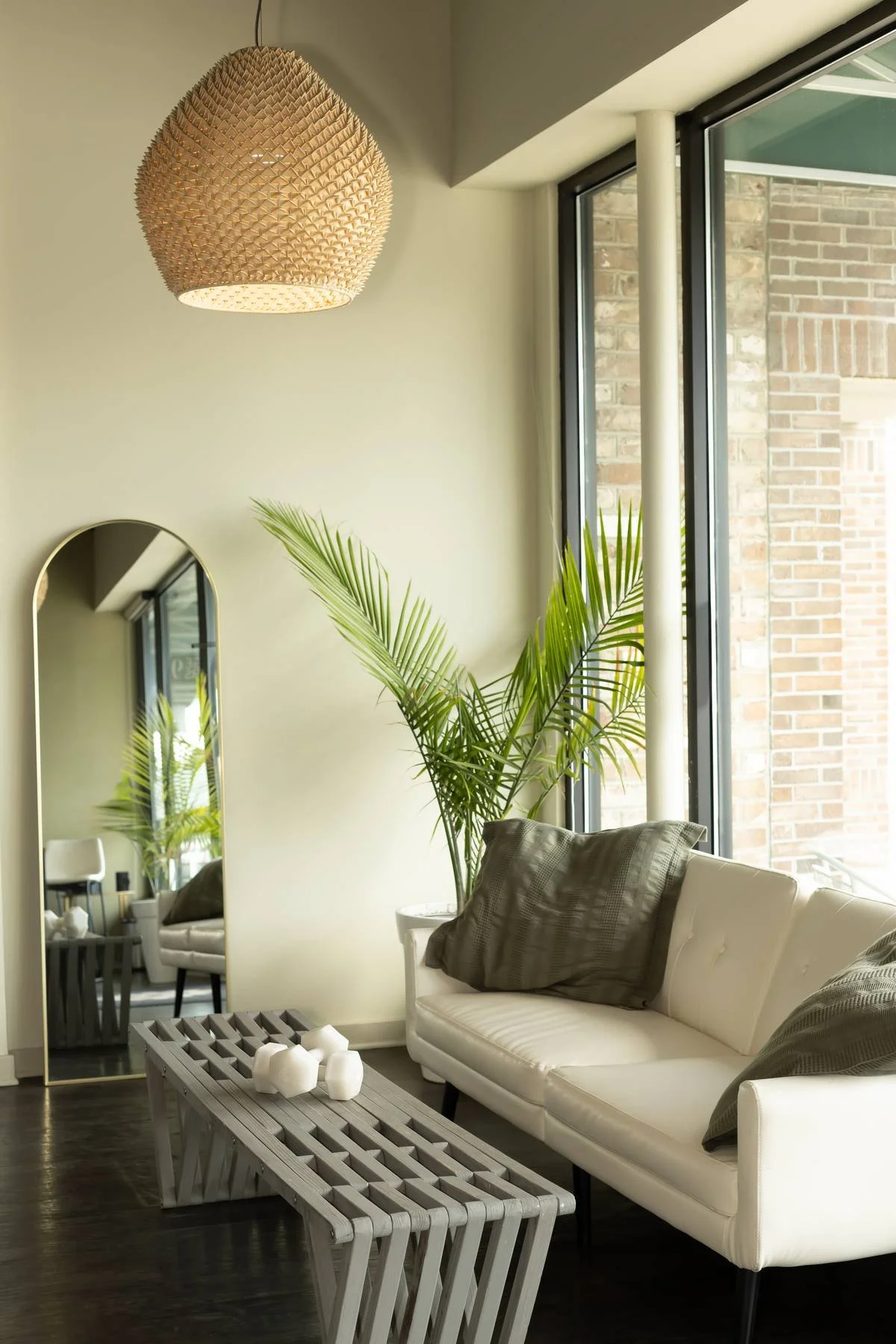 Modern living room with a white sofa, dark pillows, large window, green plant, tall mirror, woven pendant light, and wooden coffee table with candles.