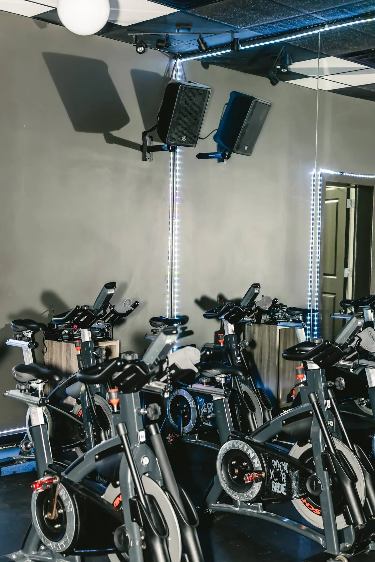Indoor cycling studio with multiple stationary bikes arranged in rows and mounted speakers on the wall.