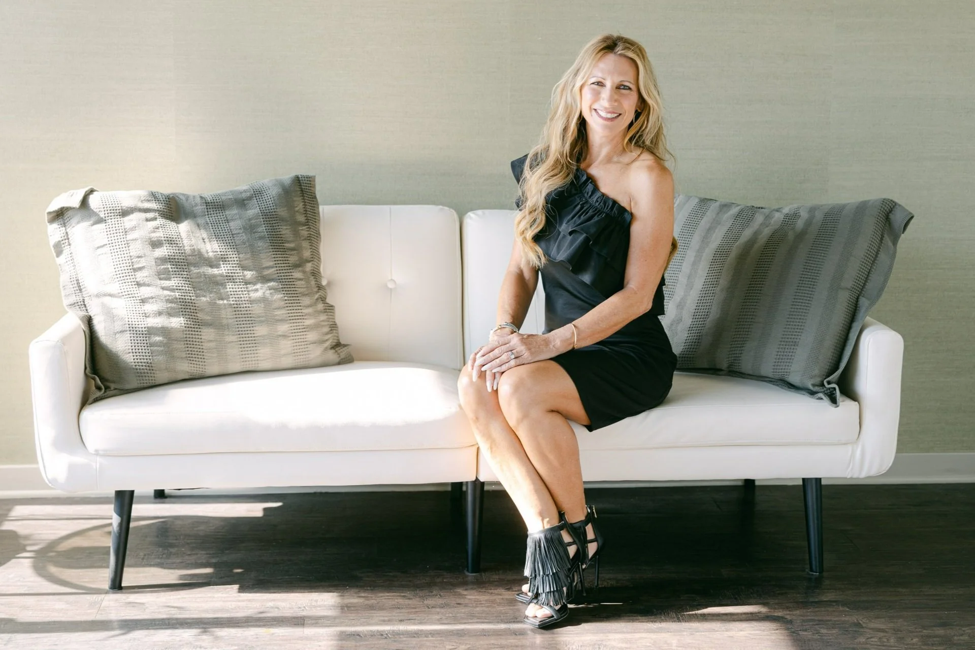 A woman with long hair sitting on a white sofa, smiling, with two gray decorative pillows on either side, against a light green wall.
