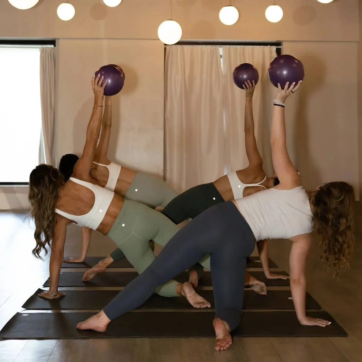 Three women participating in a yoga class, each holding a purple exercise ball overhead with one hand while in a side plank position on yoga mats in a room with beige curtains and hanging round lights.