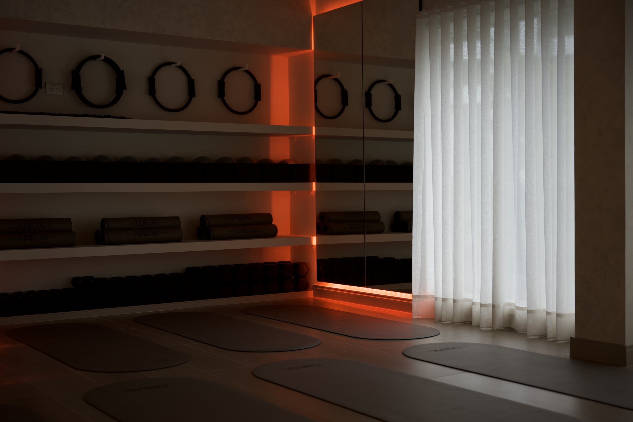 Sala de yoga con esteras en el suelo, anillos de pilates y pesas en estantes, iluminación roja y cortinas blancas.