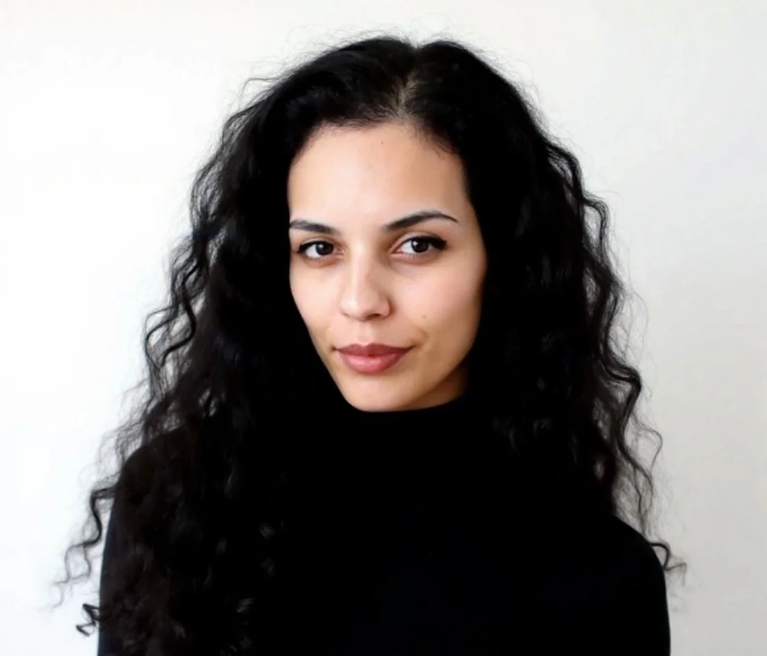 Young woman with long, curly black hair wearing a black top, standing against a plain white background.