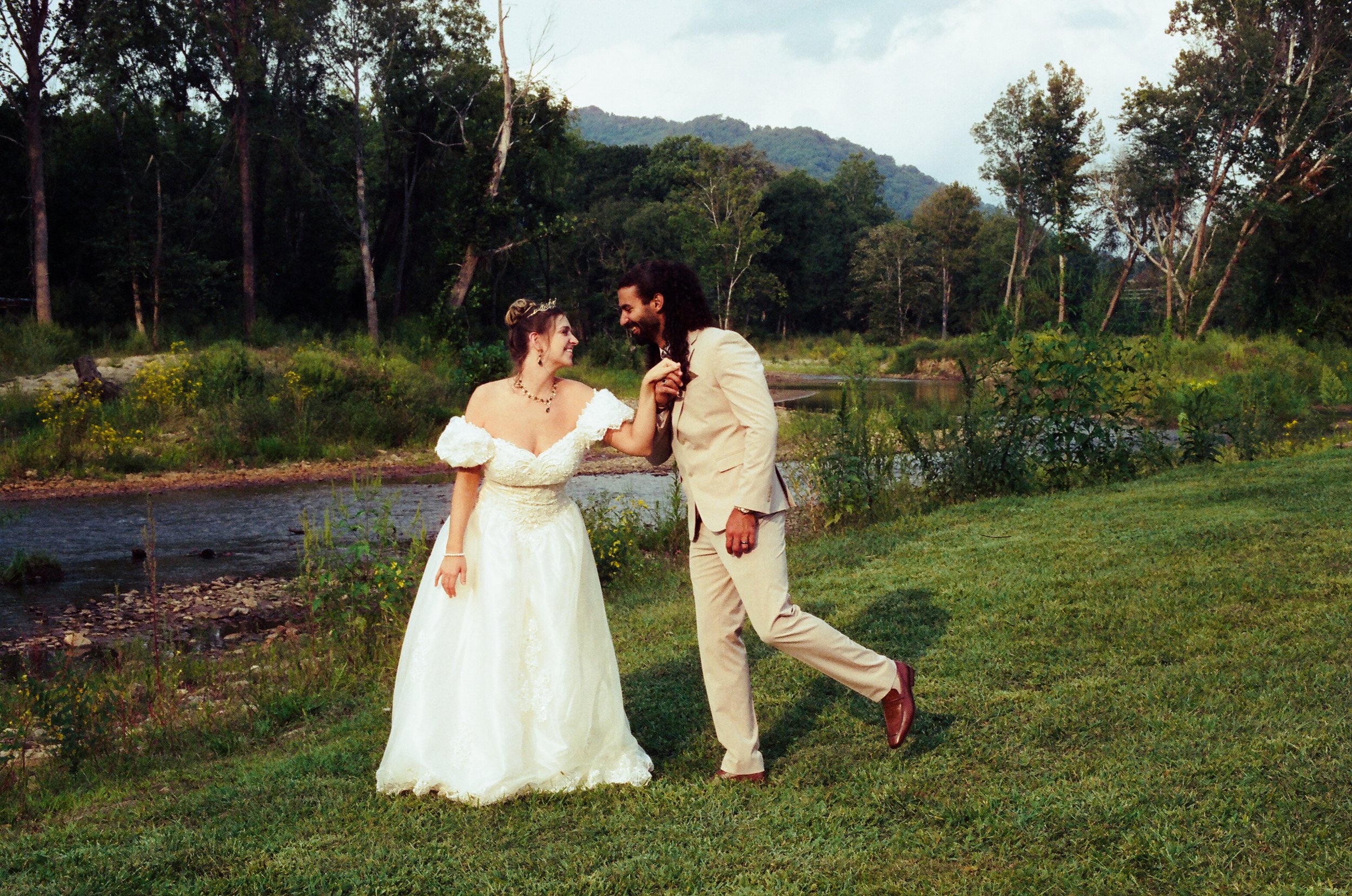 A joyful couple dressed in wedding attire standing on grass near a river with a forested area and mountains in the background.