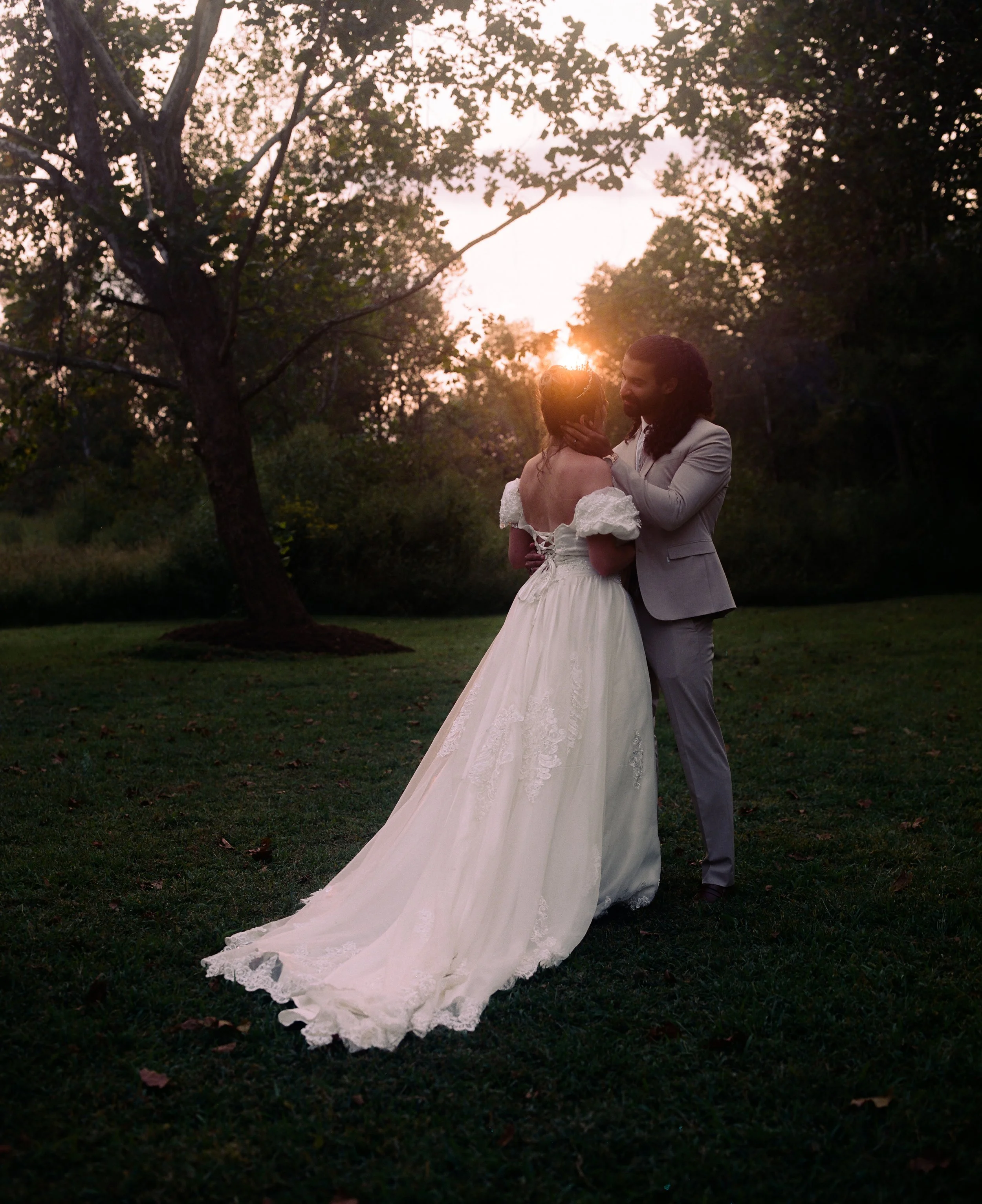 A couple in wedding attire sharing a kiss outdoors at sunset, with trees and a grassy area in the background.