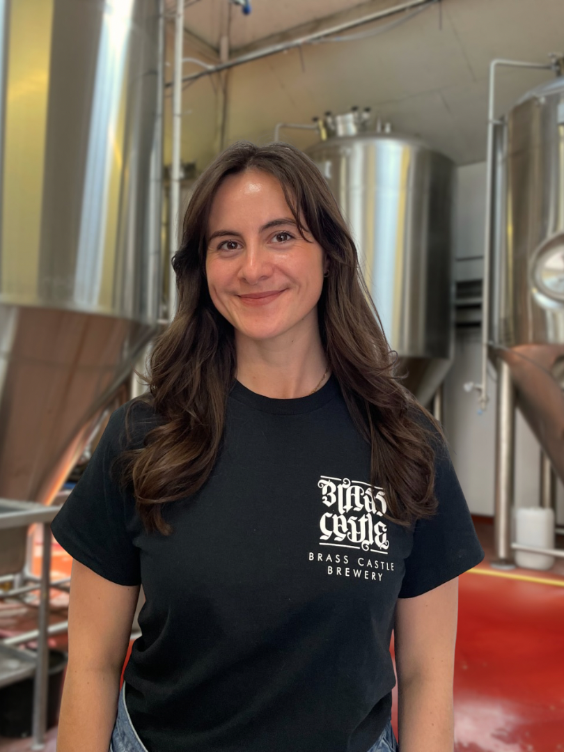 A woman with brown hair smiling, wearing a black Brass Castle Brewery t-shirt, standing in a brewery with large stainless steel brewing equipment in the background.