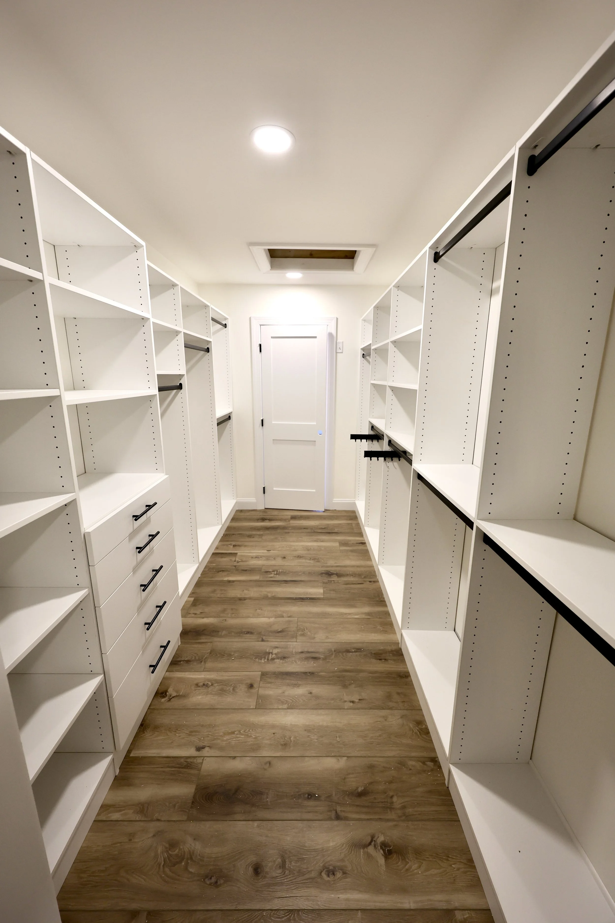 Empty walk-in closet with white shelving and black hanging rods, wood floor, ceiling lights, and a door at the end.