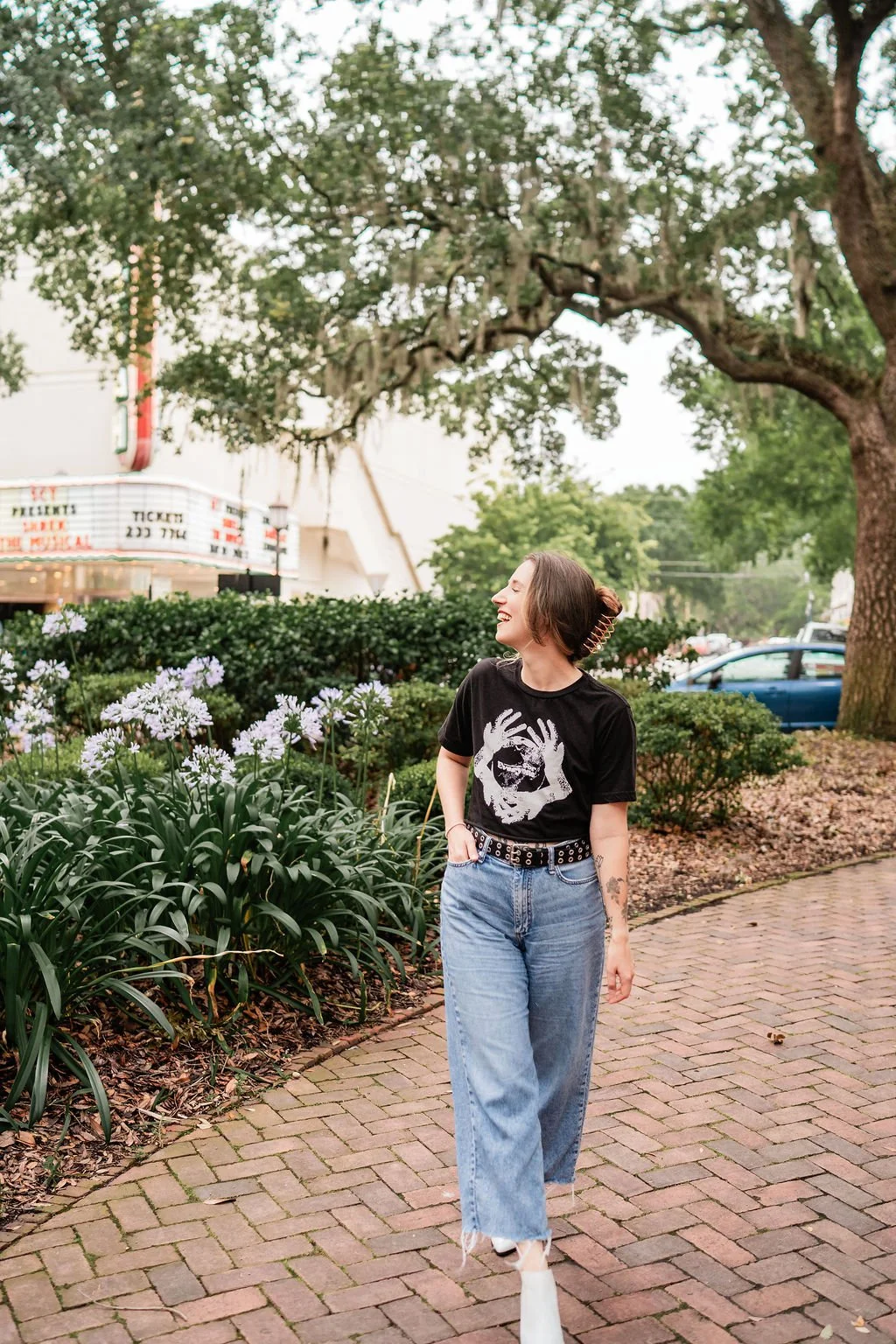 A woman walking on a brick sidewalk, smiling and looking to her right, with white flowers and green bushes on the side, large trees in the background, and a marquee sign for a theater.