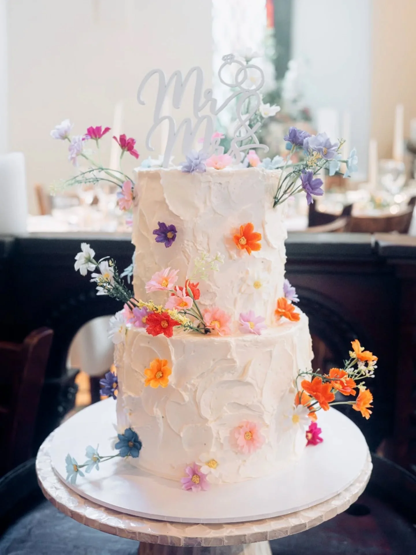 Two-tiered wedding cake with white frosting decorated with colorful edible flowers and a 'Mr & Mrs' topper with two interlocked wedding rings.
