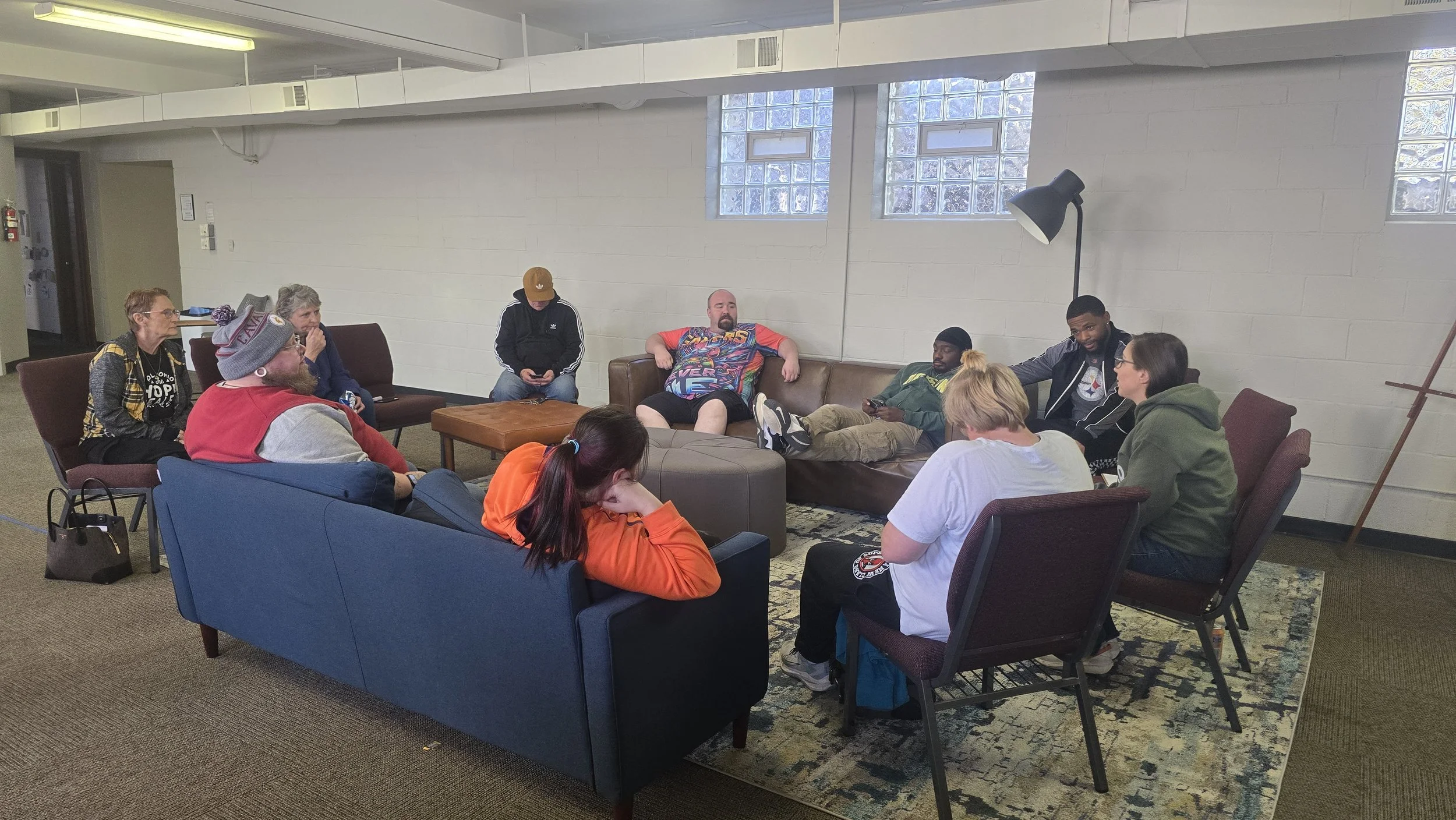 A group of people sitting in a circle in a room, some on chairs, some on couches, engaged in conversation or using phones.