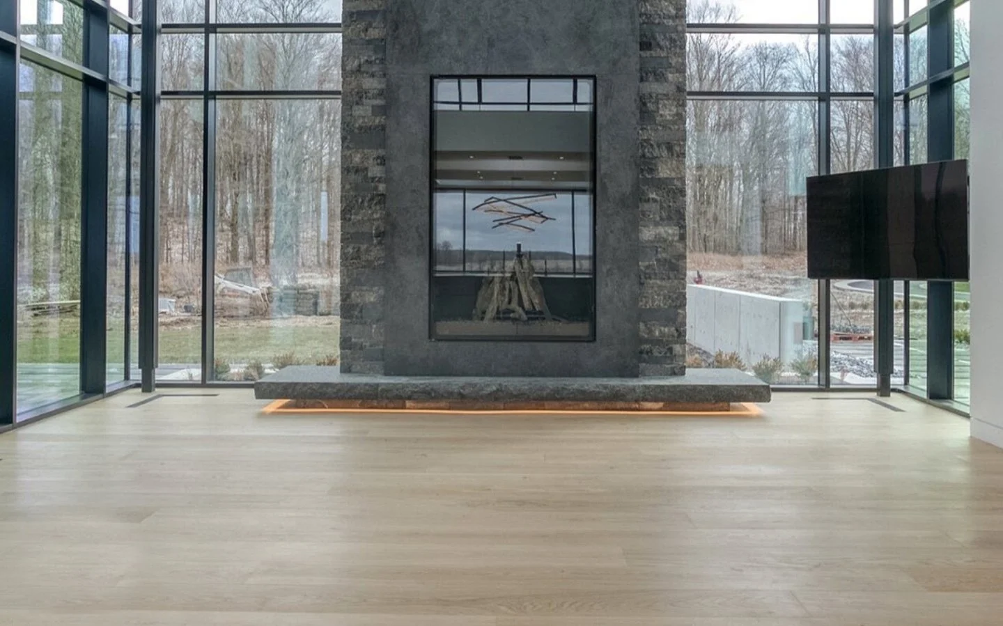 Light oak wide plank hardwood flooring in a modern glass-walled living room with a stone fireplace.