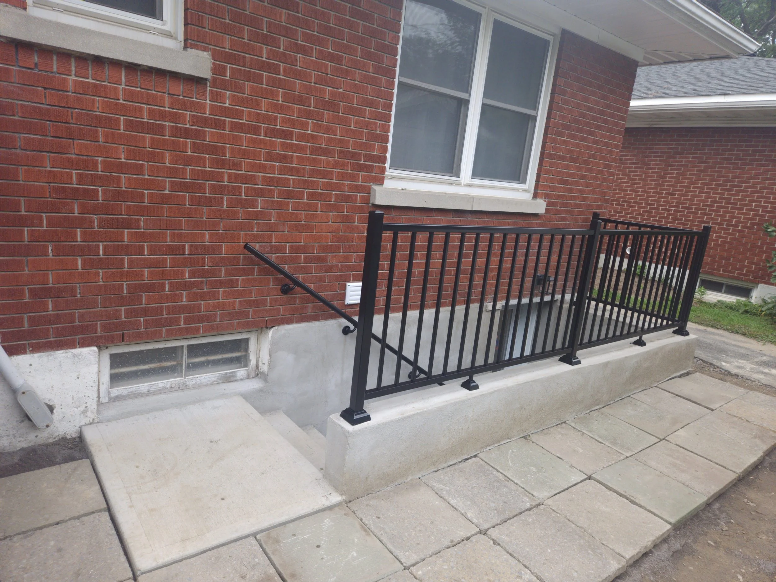 A brick house exterior with a window, a metal railing, a vent, and a concrete sidewalk.