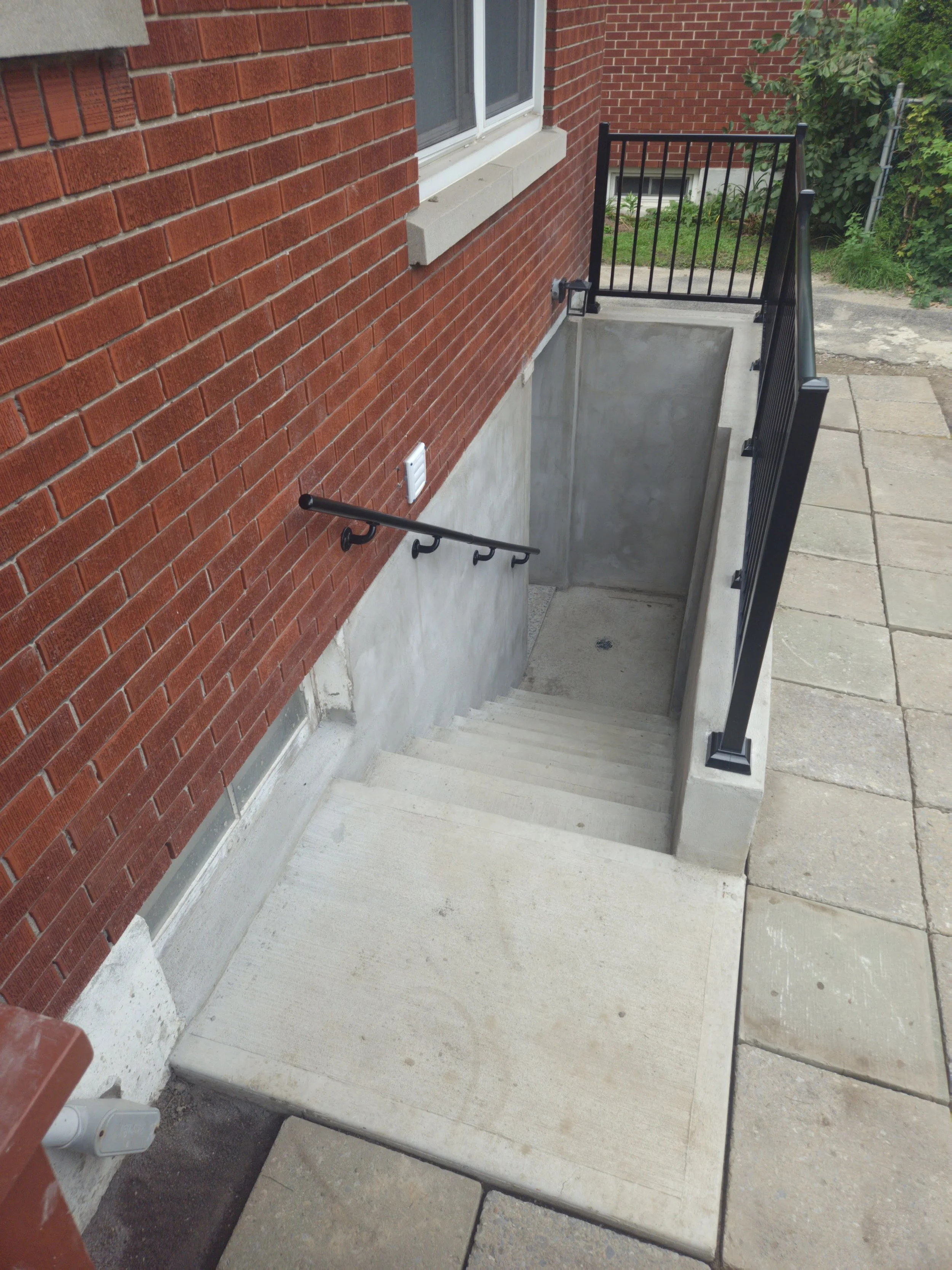 A concrete basement stairway with black metal handrails leading down to an underground entrance, adjacent to a red brick house, with some green shrubbery in the background.