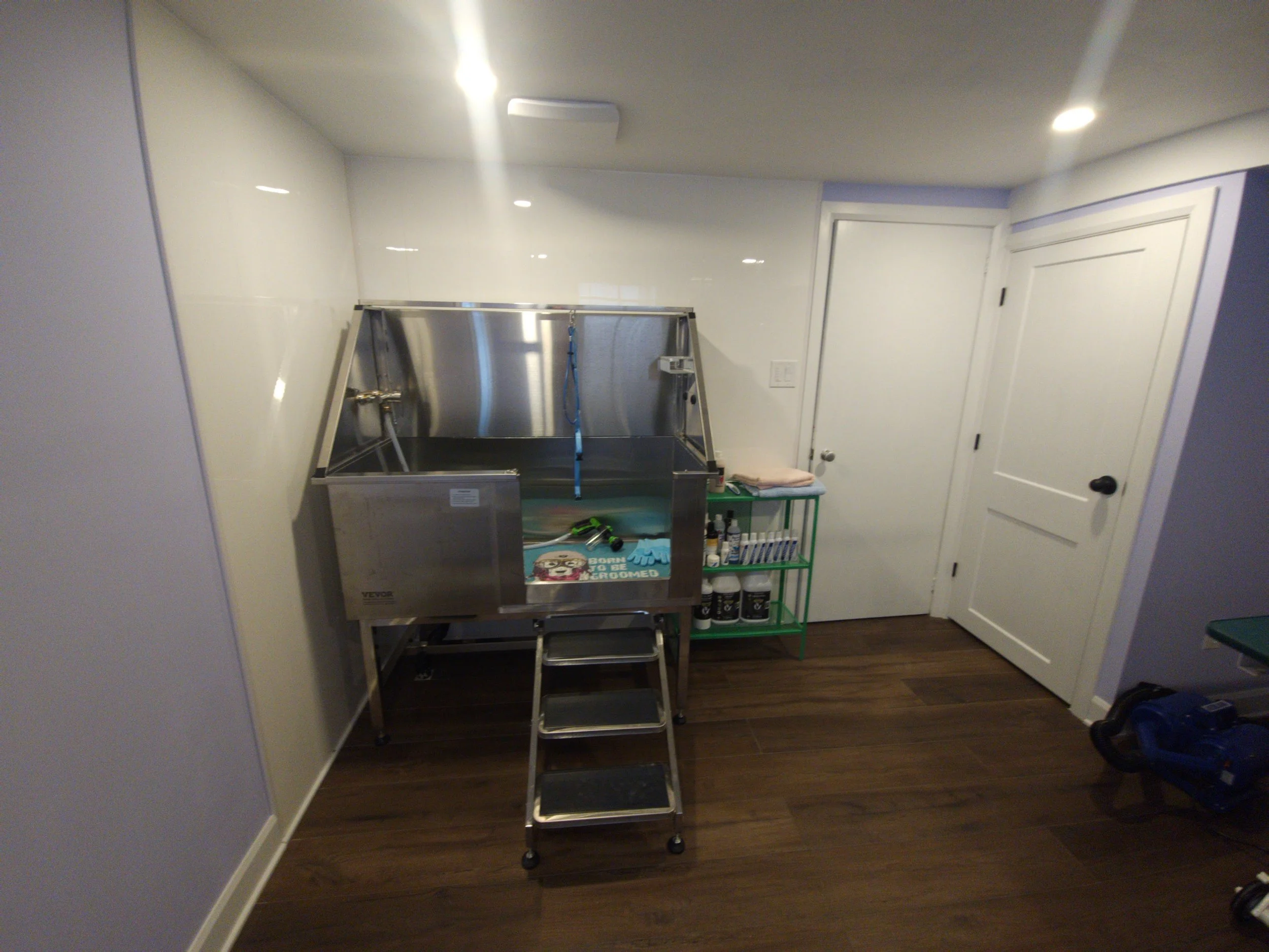 A partly assembled stainless steel pet washing or grooming station with a step ladder in front of it, placed in a room with dark wooden flooring, white walls, and a closed white door.