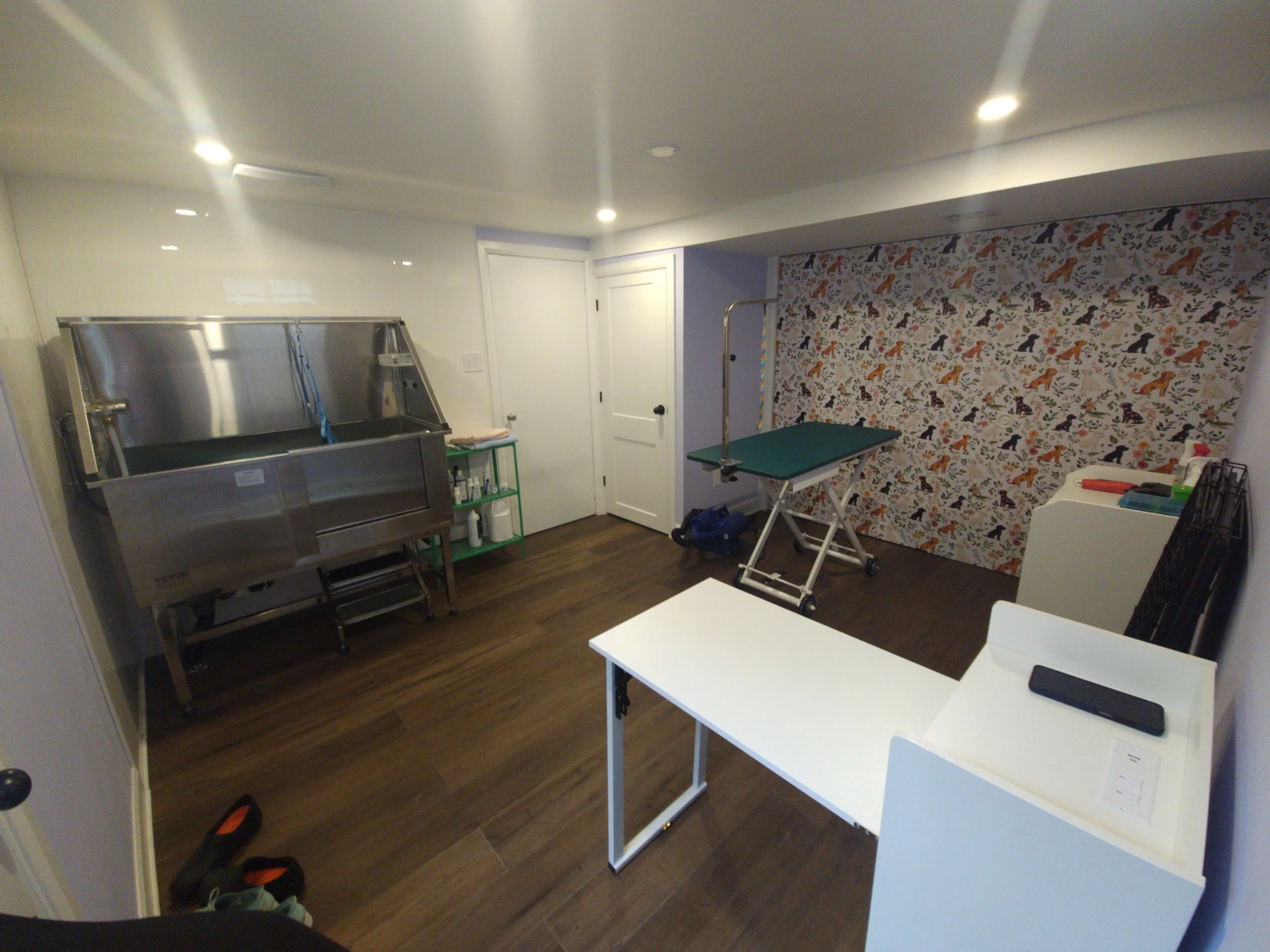 Room with white ceiling and walls, a patterned wallpaper with animals and leaves, a large metal pet grooming tub, a green trolley with cleaning supplies, an ironing table, and white shelving units. Shoes are on the dark wooden floor. Three ceiling li