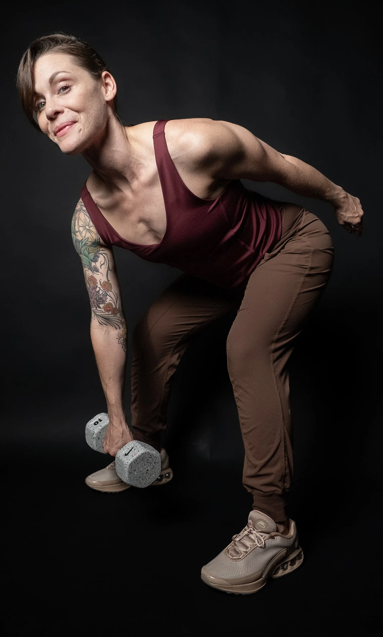 A woman with short brown hair, tattoos on her left arm, wearing a maroon tank top, brown pants, and beige sneakers, exercises with a dumbbell against a black background.