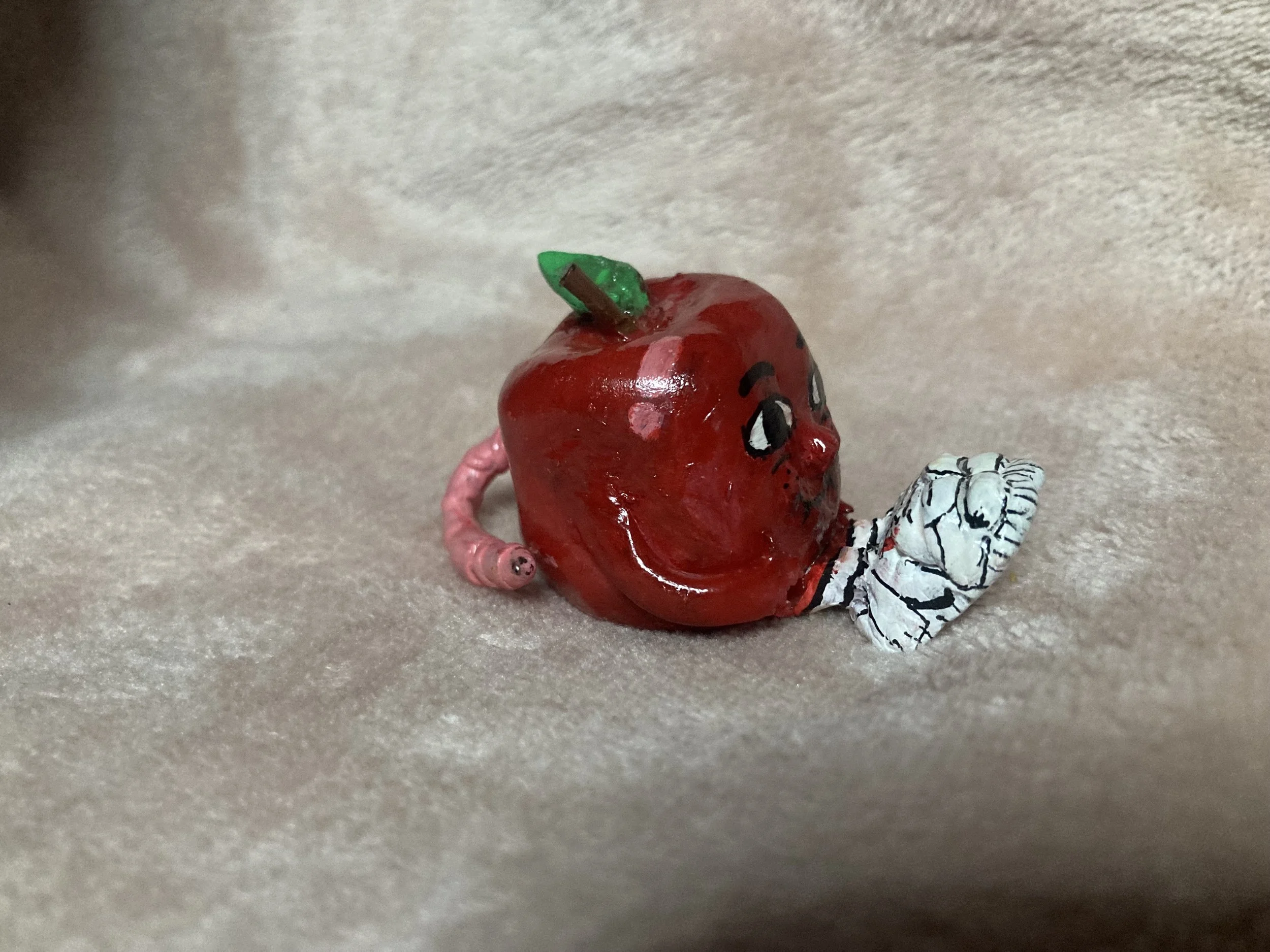 A small, red, cartoonish apple figure with a painted face, next to a white and black patterned sock, all on a soft beige fabric background.