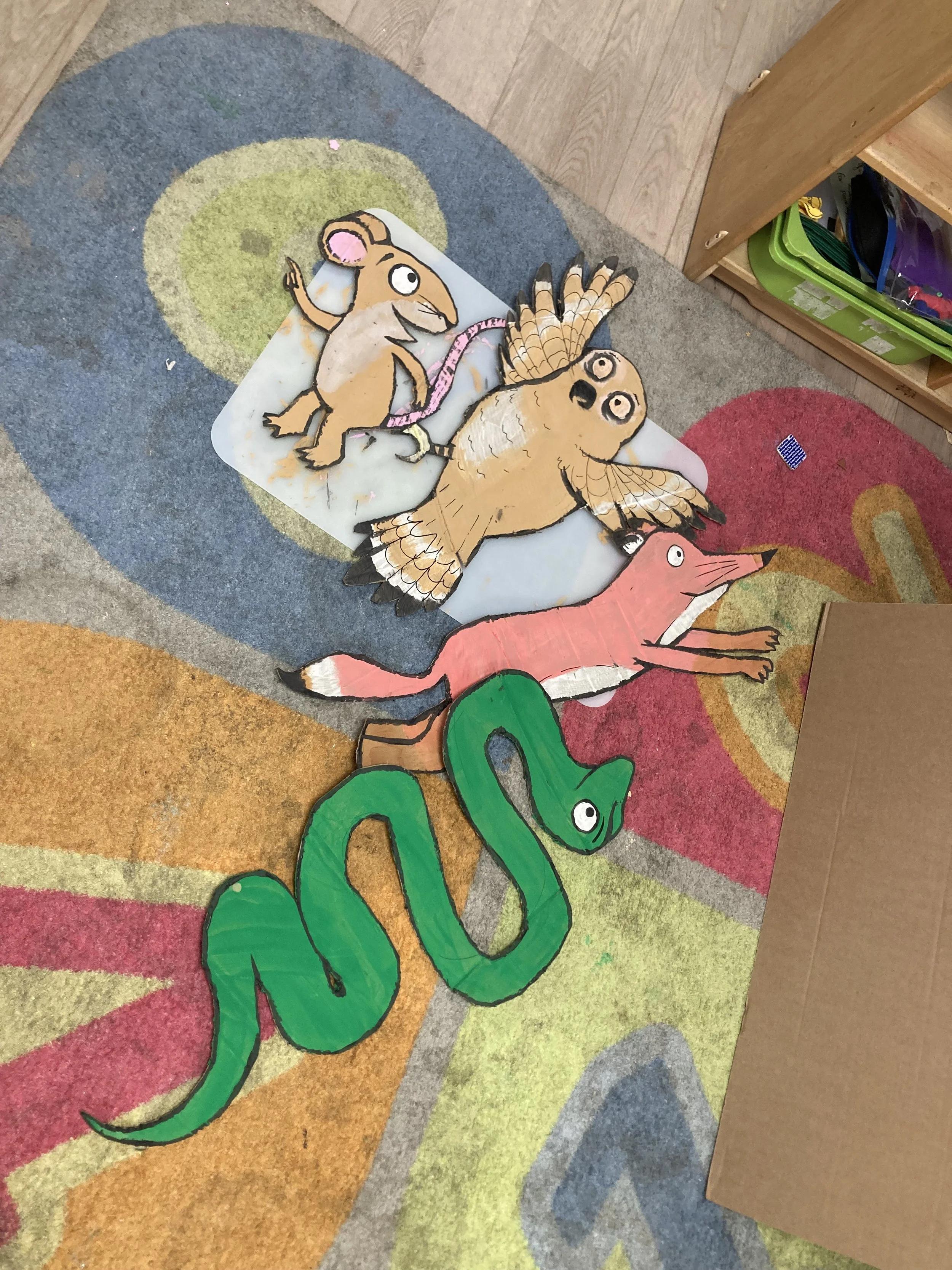Colorful cartoon animals on a rainbow-colored carpet, including a green snake, a pink fox, an owl, and a mouse with a pink ribbon.