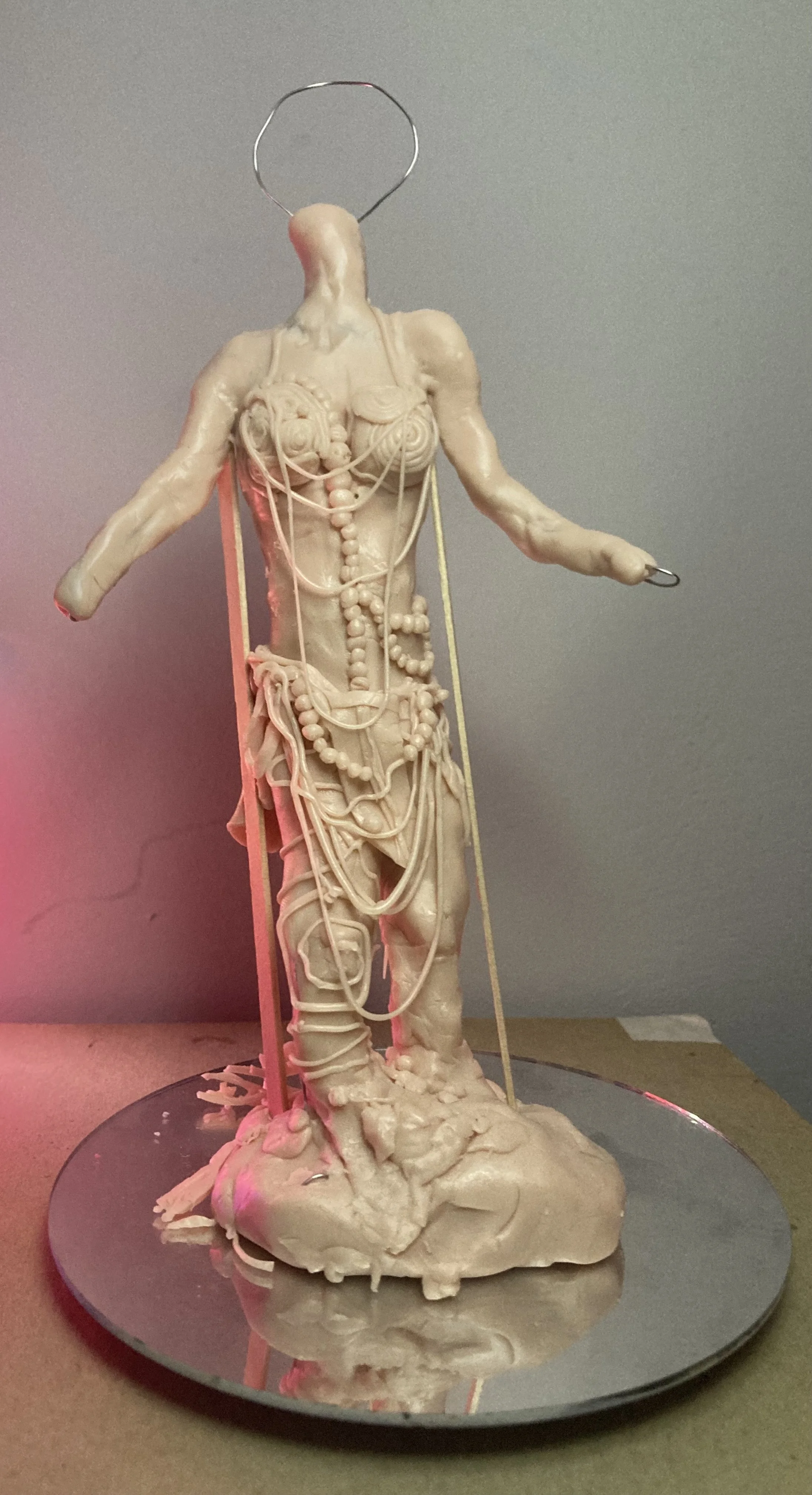 An abstract sculpture resembling a human figure made from various materials including string, beads, and antlers, standing on a reflective surface with a neutral background.