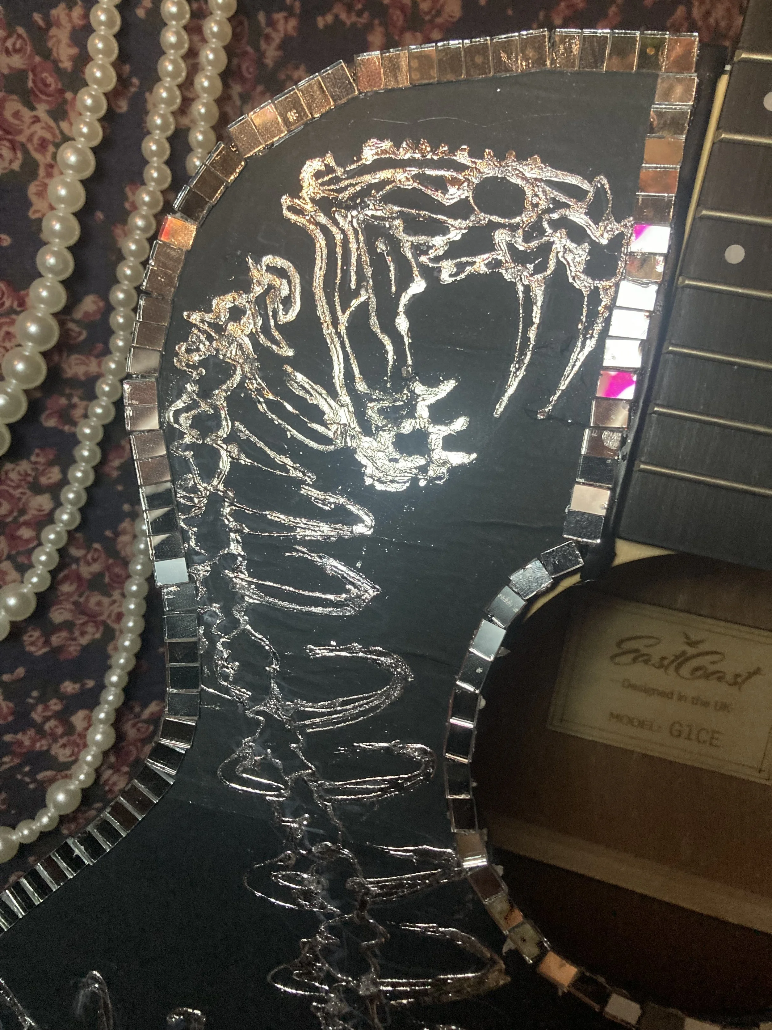 Decorative black mirror with silver mosaic border, reflecting light and surroundings, with a pearl necklace nearby and a wooden box labeled 'East Coast' and 'Model G1CE' in the background.