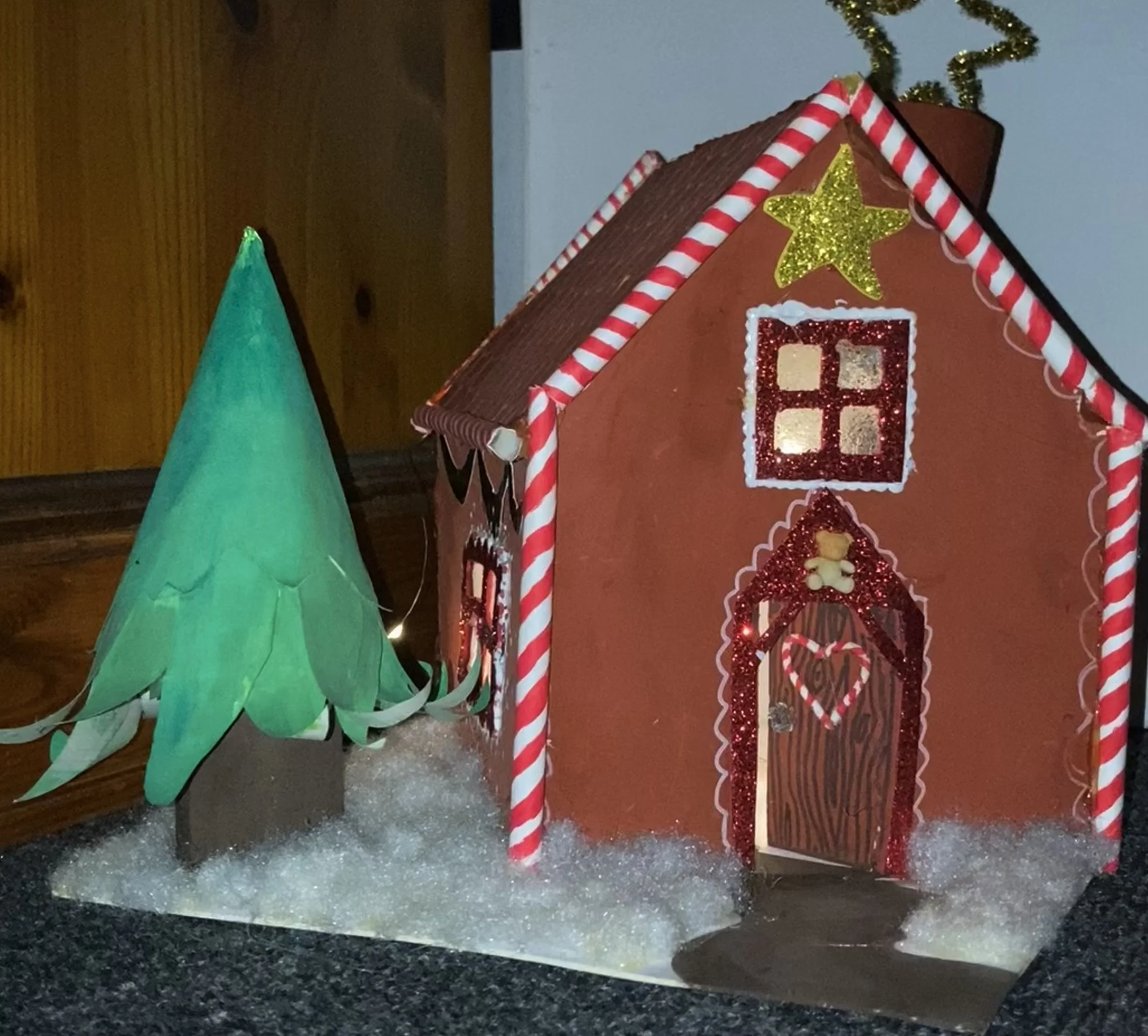 A gingerbread house decorated with red and white striped candy canes, a golden star, and red glitter accents, placed next to a green paper Christmas tree, on a snowy base.