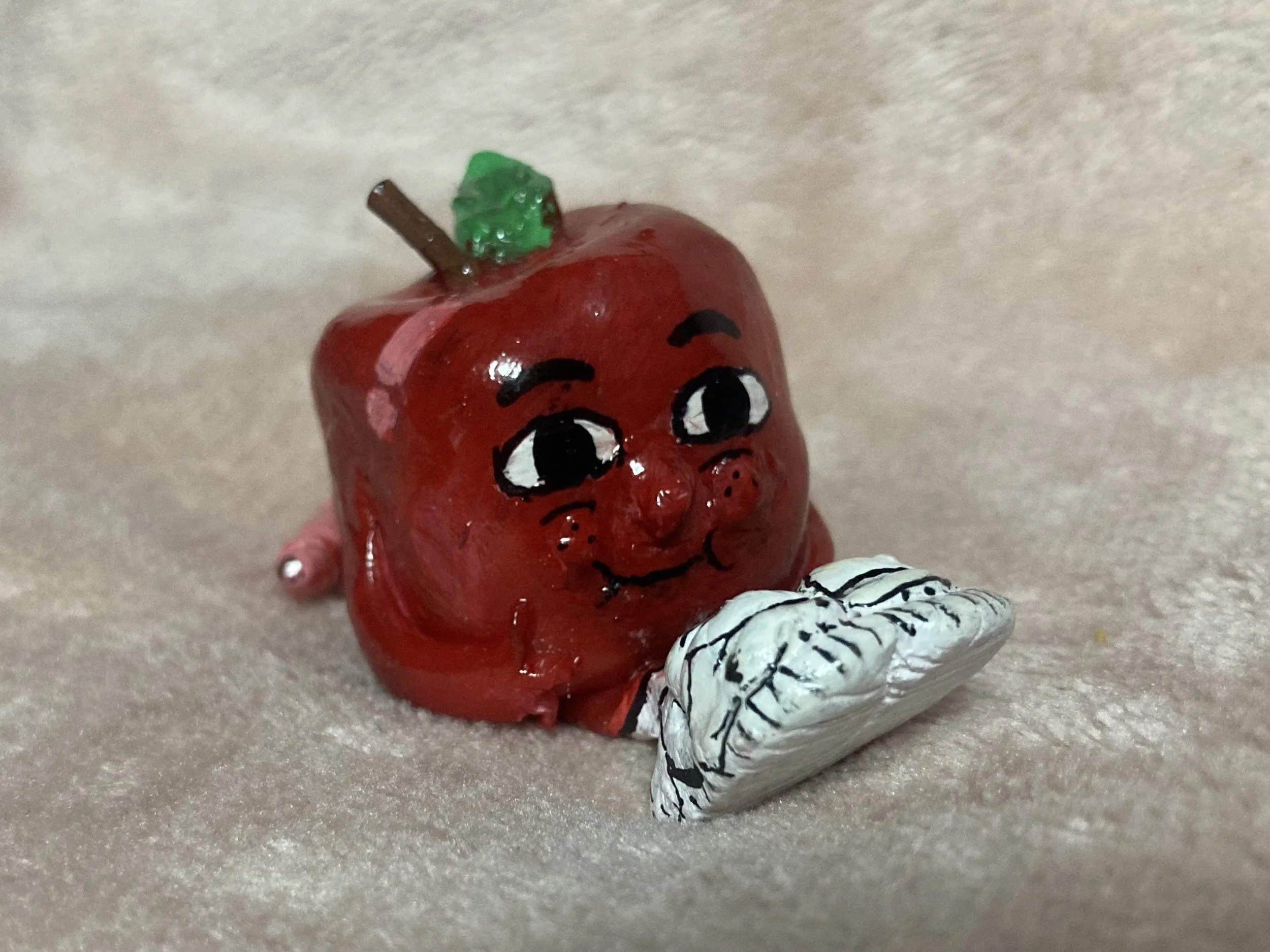 A small, red, apple-shaped figurine with a smiling face, big eyes, and a small leaf and stem on top. It is holding a white, textured object resembling a leaf or a book.