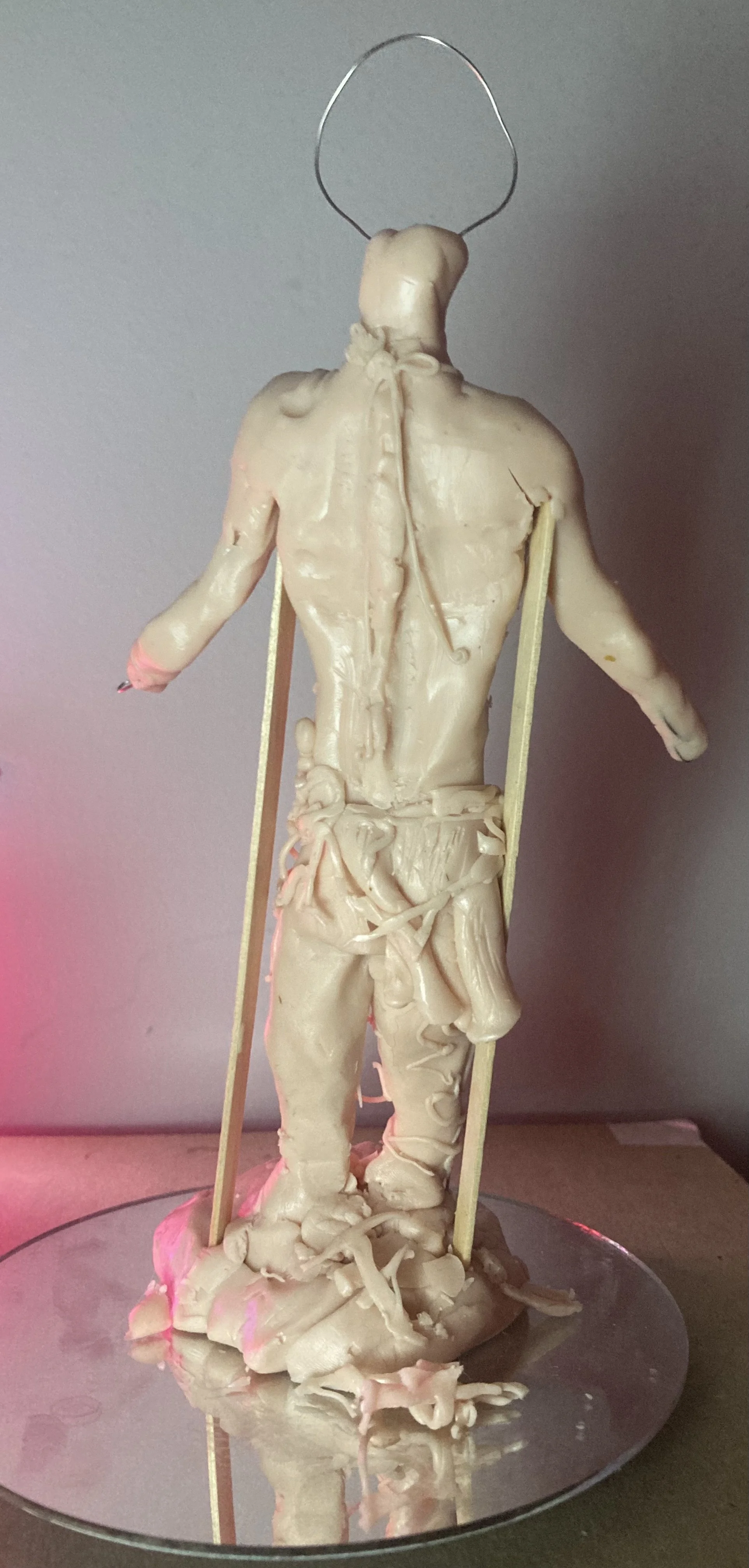 A sculpture of a human figure with a head made of wire, no facial features, and body parts that are incomplete or melted, mounted on a reflective surface.