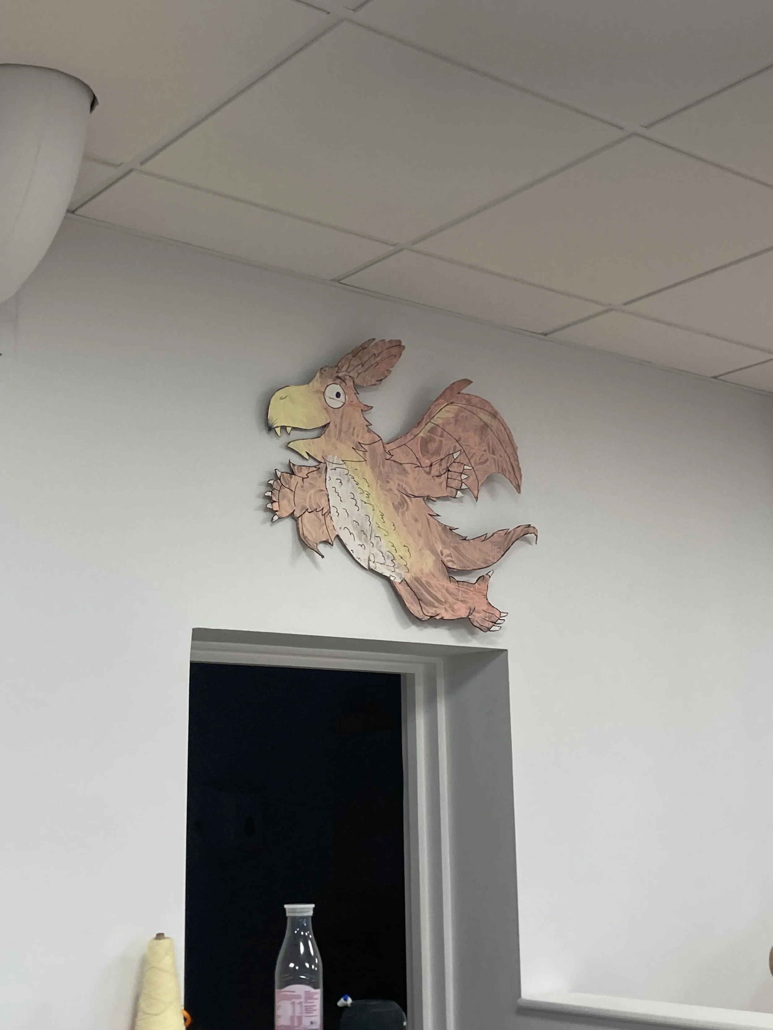 A colorful dragon art piece made of paper or similar material, mounted on a white wall above an open doorway in an indoor setting.