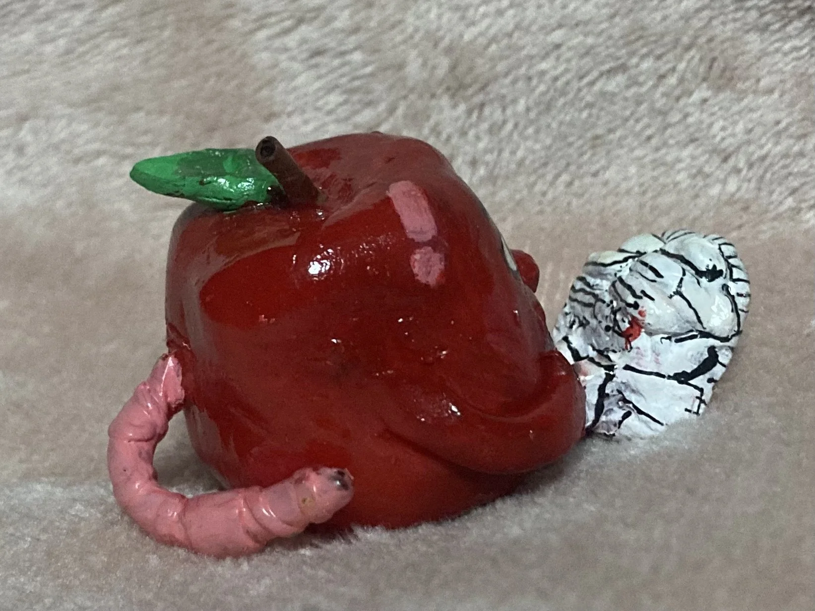 A model of an apple made from various materials, with a green leaf, a brown stem, a pink worm, and a black and white patterned serpent wrapped around it, lying on a soft beige surface.