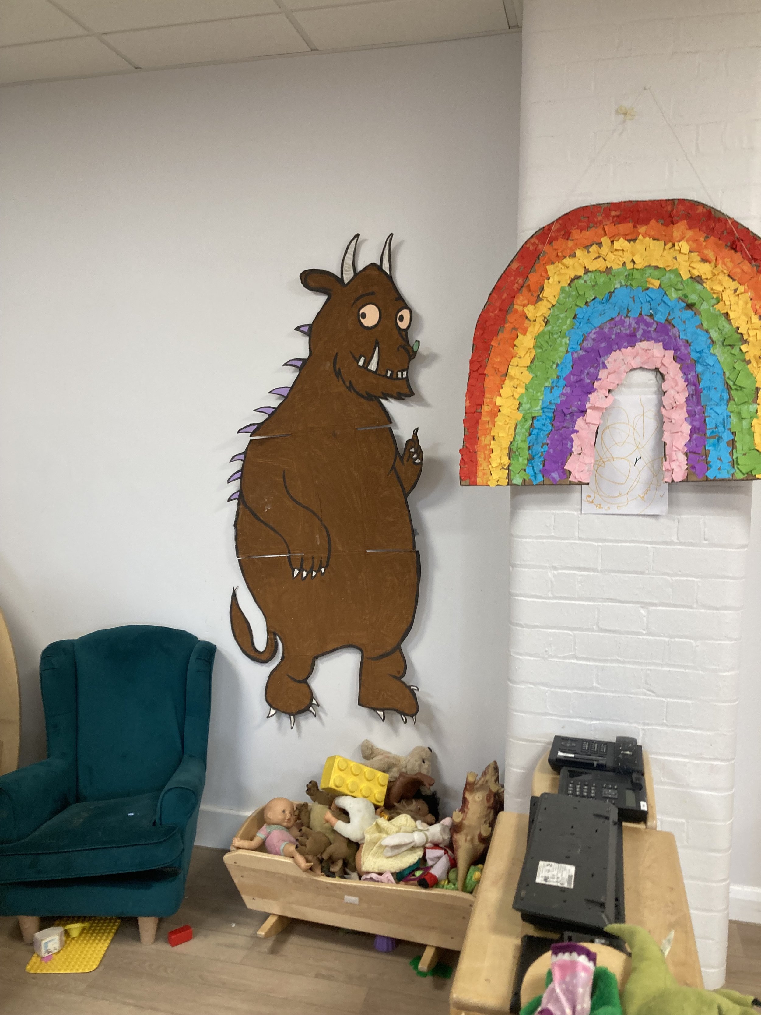 Children's play area with a large cartoon monster wall decoration, a rainbow made of tissue paper, a green armchair, and a box of toys filled with stuffed animals and dolls.