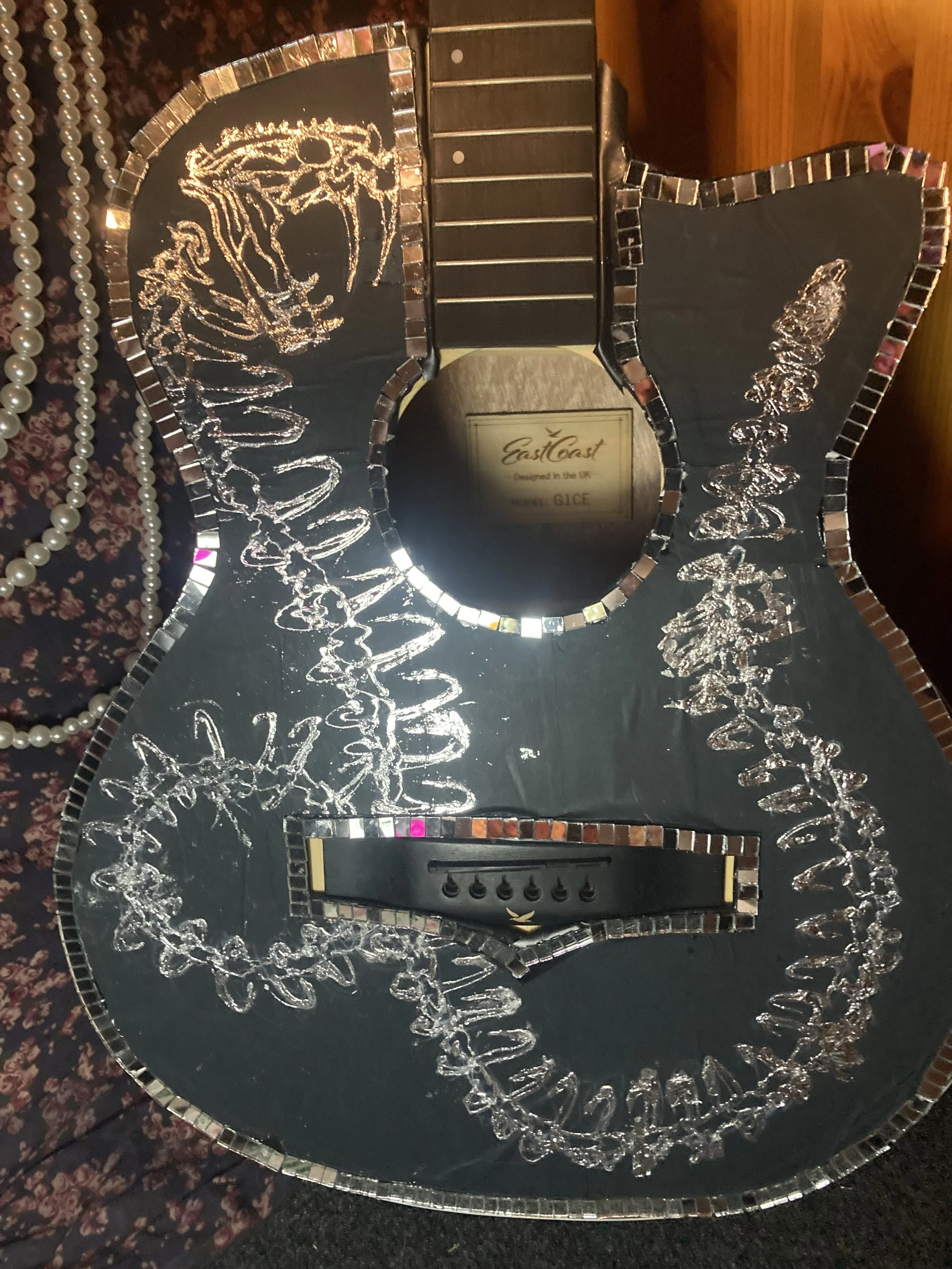 Decorative black guitar-shaped object with silver and pink mosaic edges, featuring silver engraved Russian text and skull and floral designs, and a control panel at the bottom.