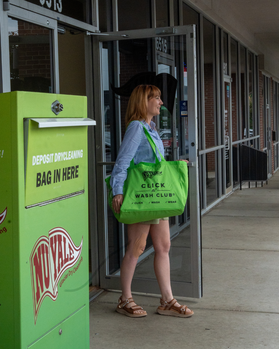 A woman standing outside a dry cleaning business, holding a large green bag, wearing a striped shirt, shorts, and sandals, with a deposit dry cleaning bag in her right hand.