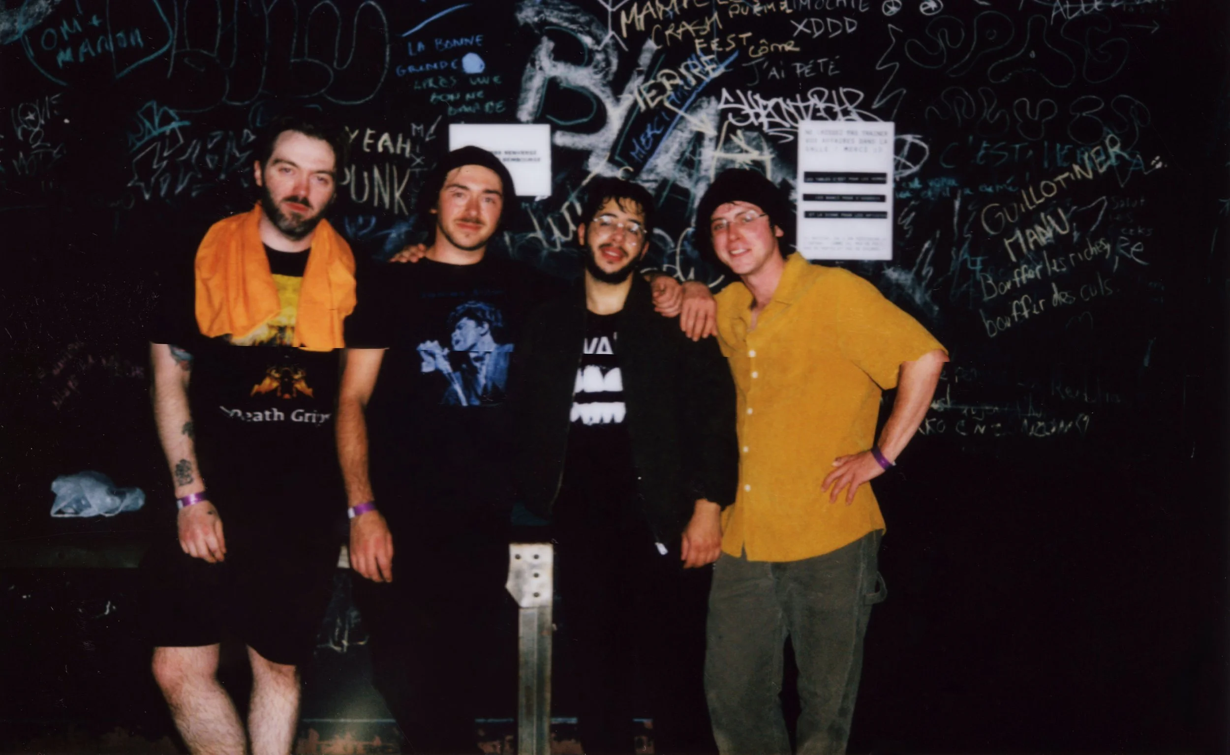 Four people stand side by side in front of a black wall covered in graffiti-style writing; their faces are blurred, and they wear casual clothes including band T‑shirts, shorts, jeans, and a yellow short-sleeve shirt in a dimly lit, indoor setting.