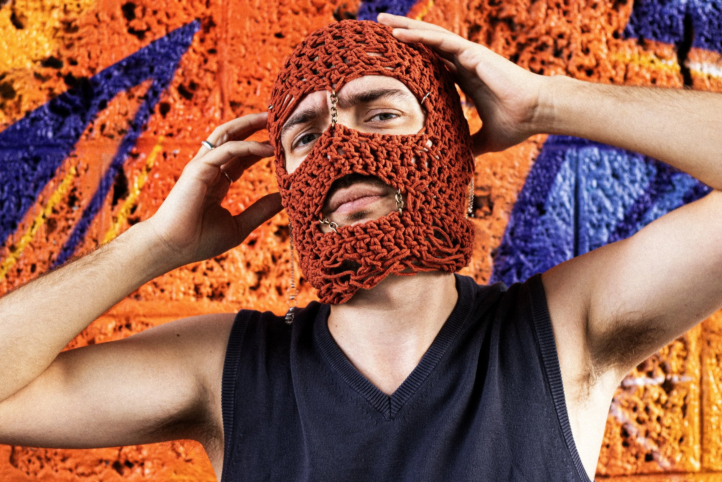 French anti‑folk artist wearing a knitted mask and sleeveless top, adjusting the mask in front of a bright orange and purple graffiti wall, photographed for a NOIZE Magazine alt‑folk artist interview by Adele Monty.