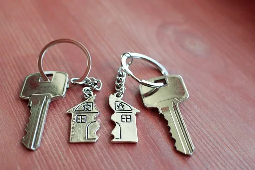 Deux clés et deux porte-clés en forme de maisons et de clés, posés sur une surface en bois.