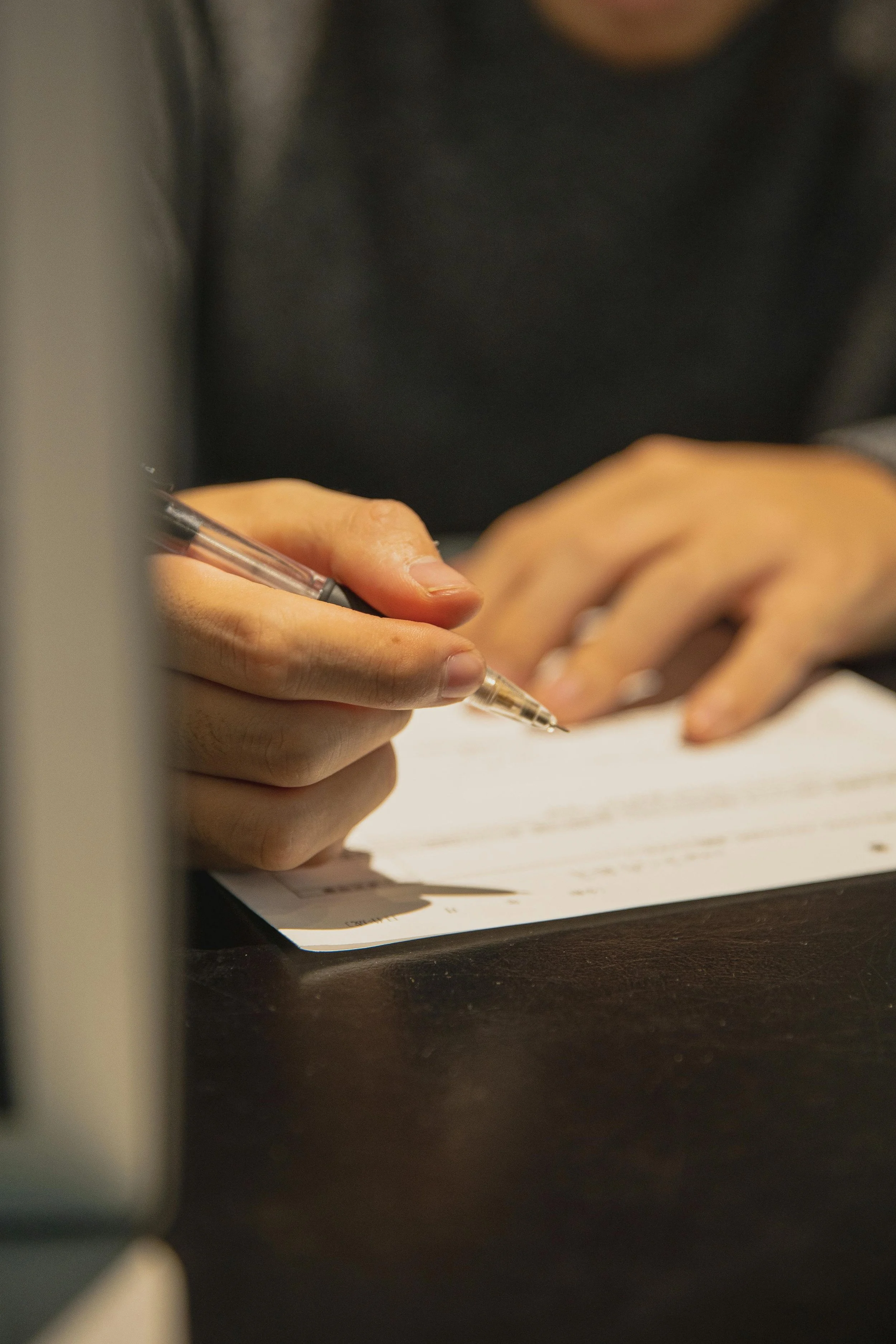 Une personne écrit avec un stylo sur une feuille de papier posée sur une table en bois, vêtue d'un pull gris foncé.