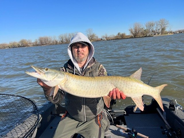 Man holding a large fish on a boat, with a lake in the background.