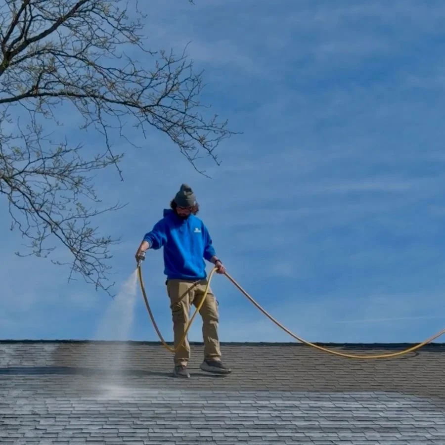 Person in a blue hoodie pressure washing a roof on a clear day.
