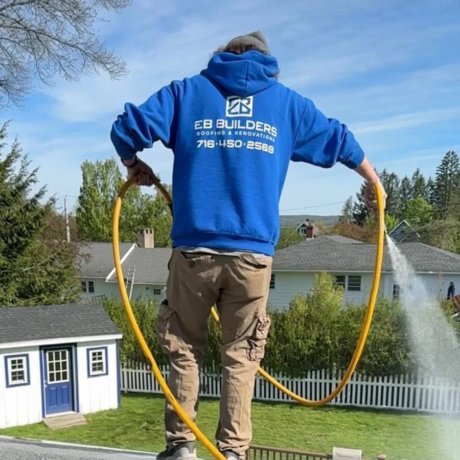 Person standing on a roof, holding a garden hose, spraying water, wearing a blue hoodie with company logo, tan cargo pants, and a gray beanie, with a backyard, trees, and houses in the background.