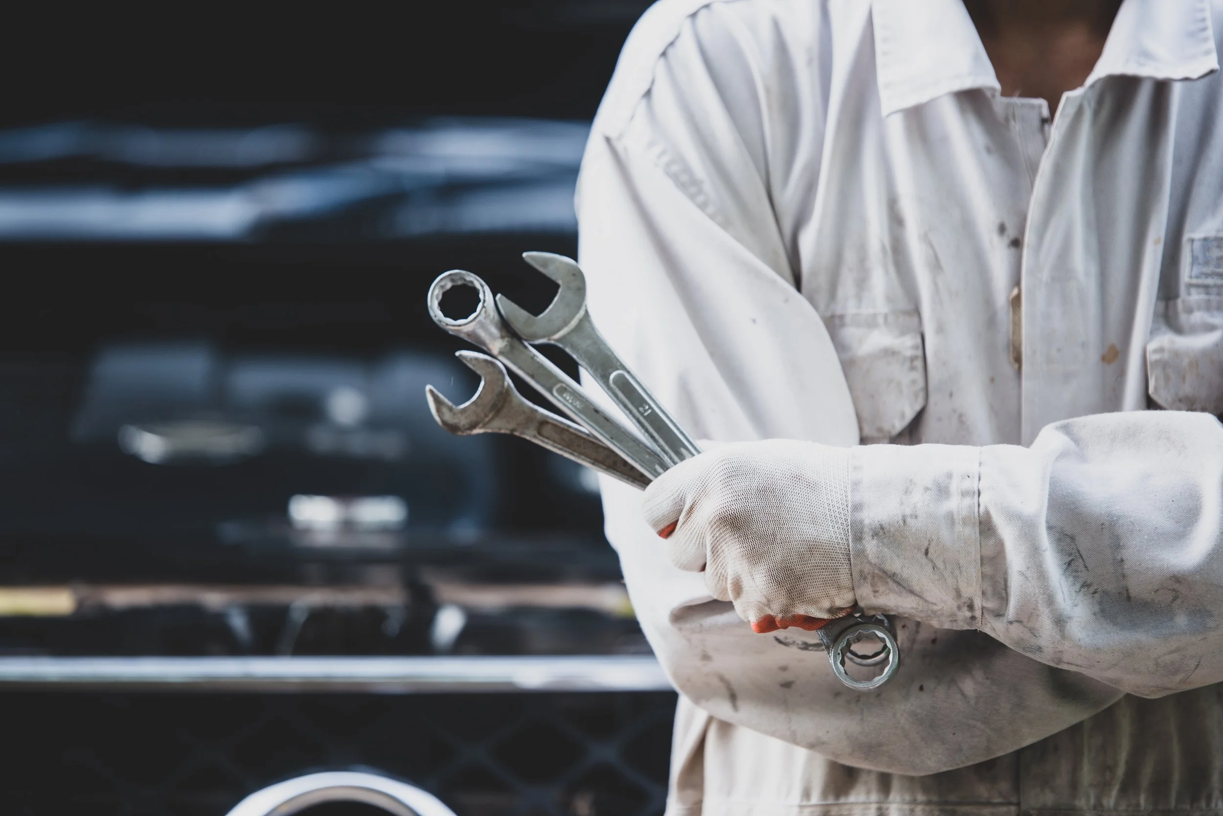 A person wearing a white mechanic uniform and gloves holding a set of wrenches in front of a car.