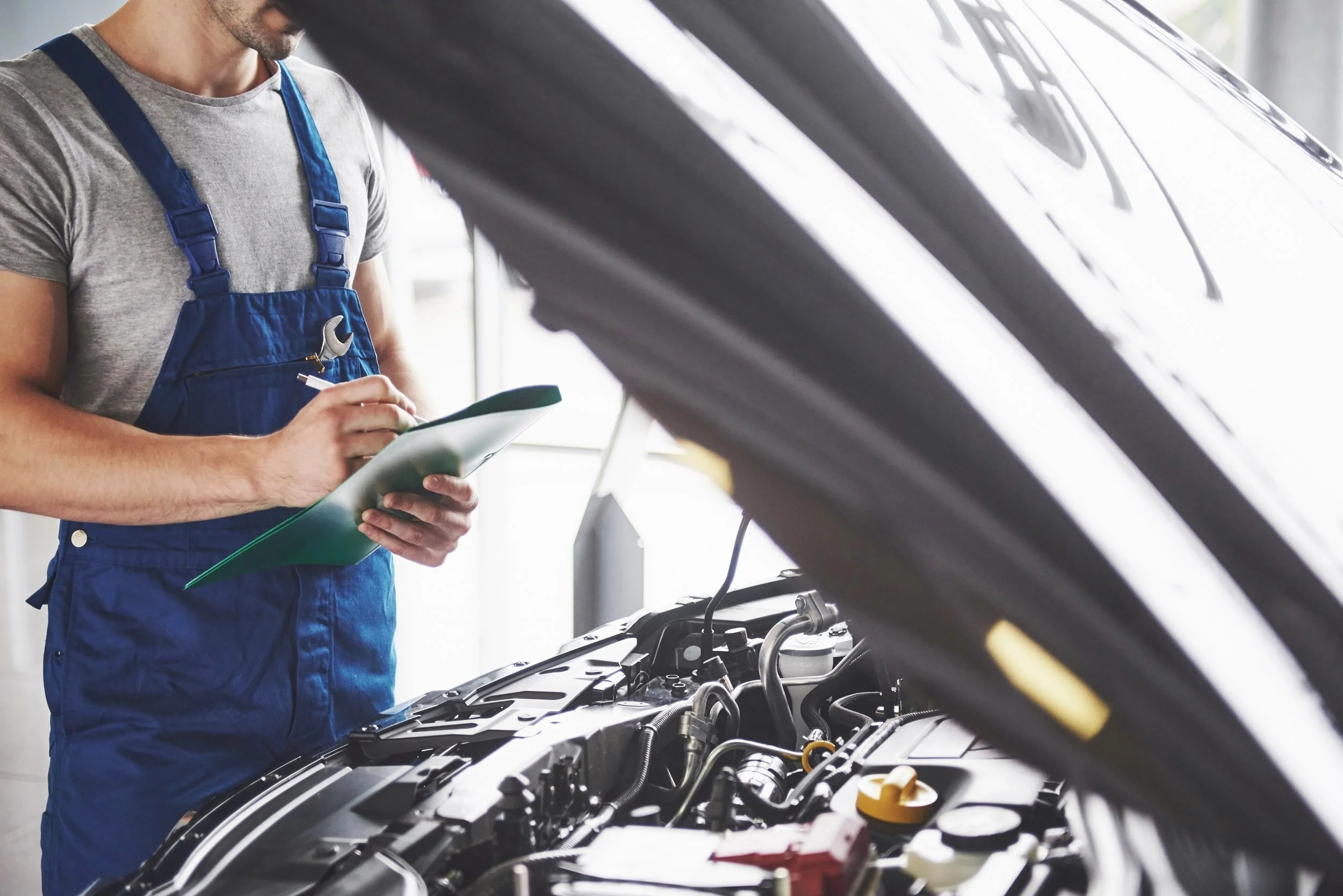 A mechanic in a gray t-shirt and blue overalls is inspecting a car engine while holding a clipboard.
